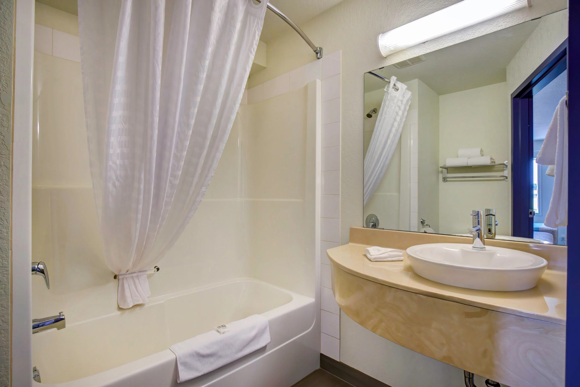 Bathroom with white tub, shower curtain, vanity sink, and large mirror under bright lighting.