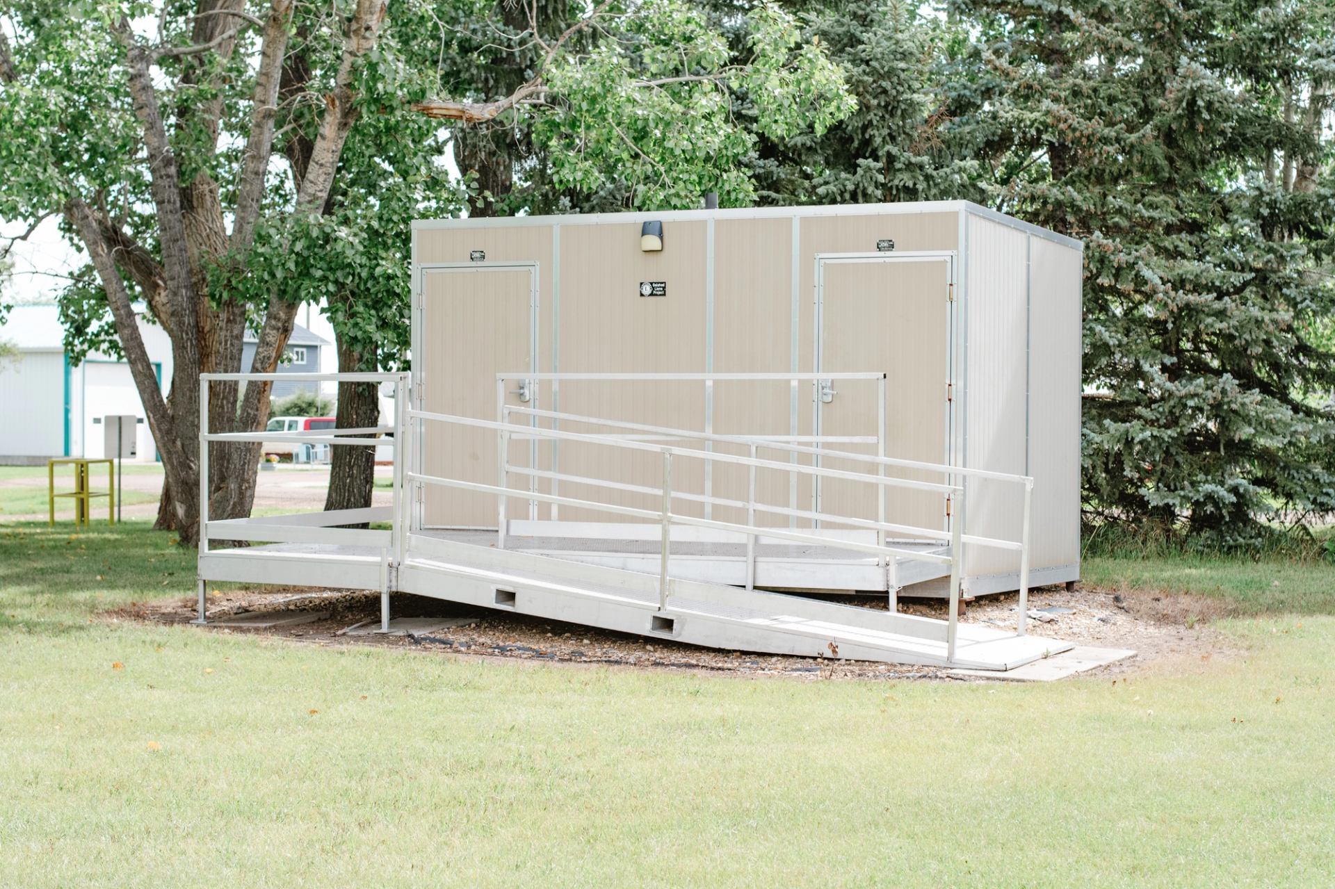 Portable beige washroom building with metal accessibility ramp, surrounded by trees and grass.