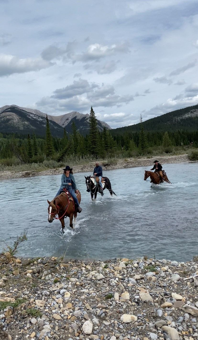 Riders on horseback crossing a shallow river with mountains nearby.