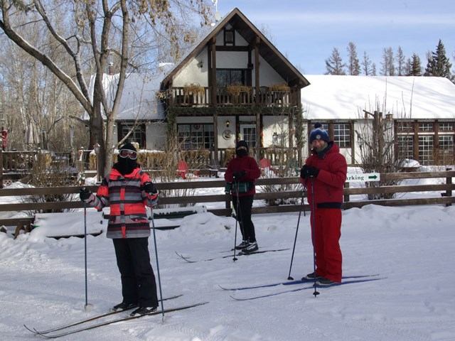 Skiers on snowy trail near lodge with winter trees and cross-country gear.
