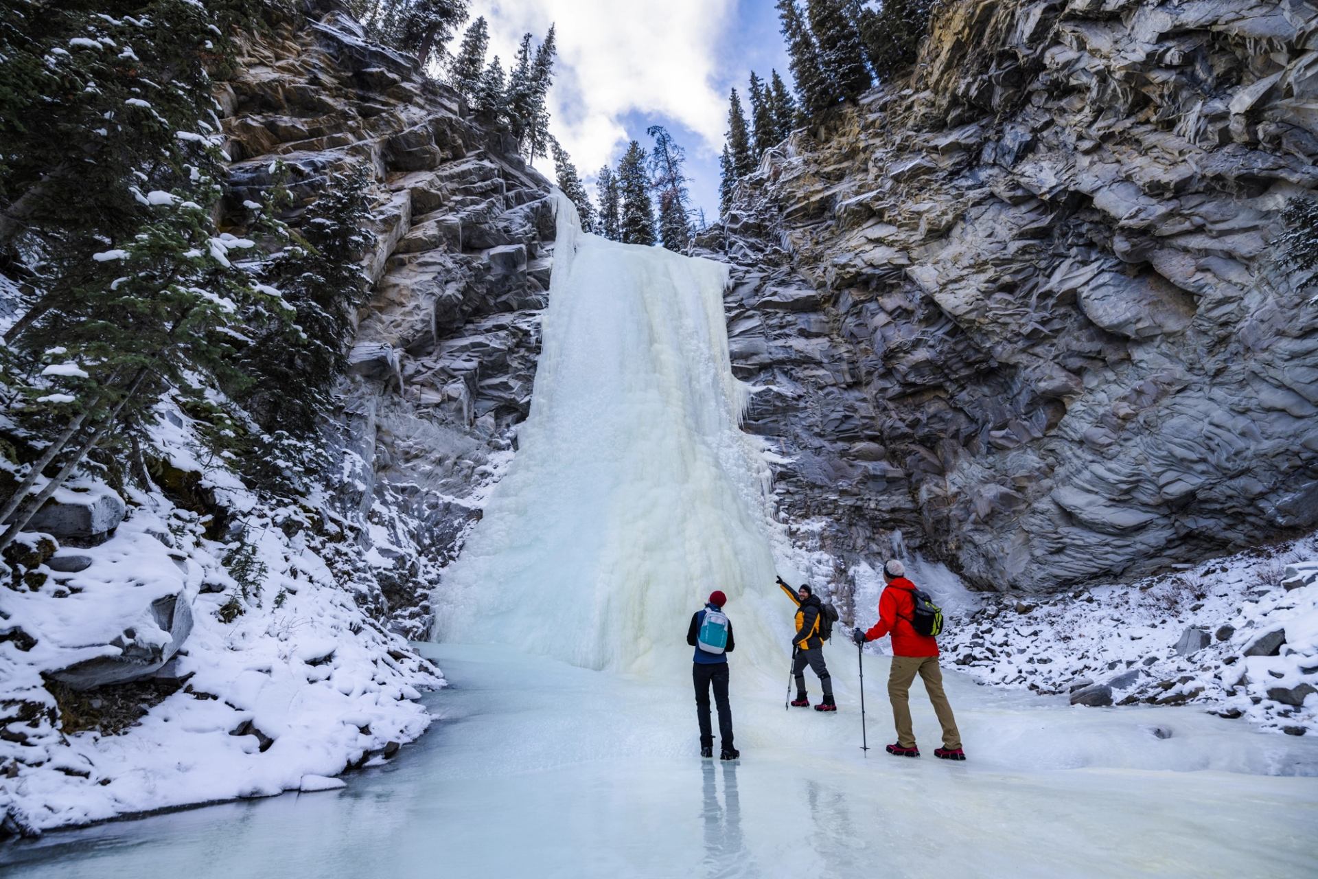 Three people stand on a frozen river below a towering ice waterfall, framed by rocky canyon walls and snow‑dusted trees.