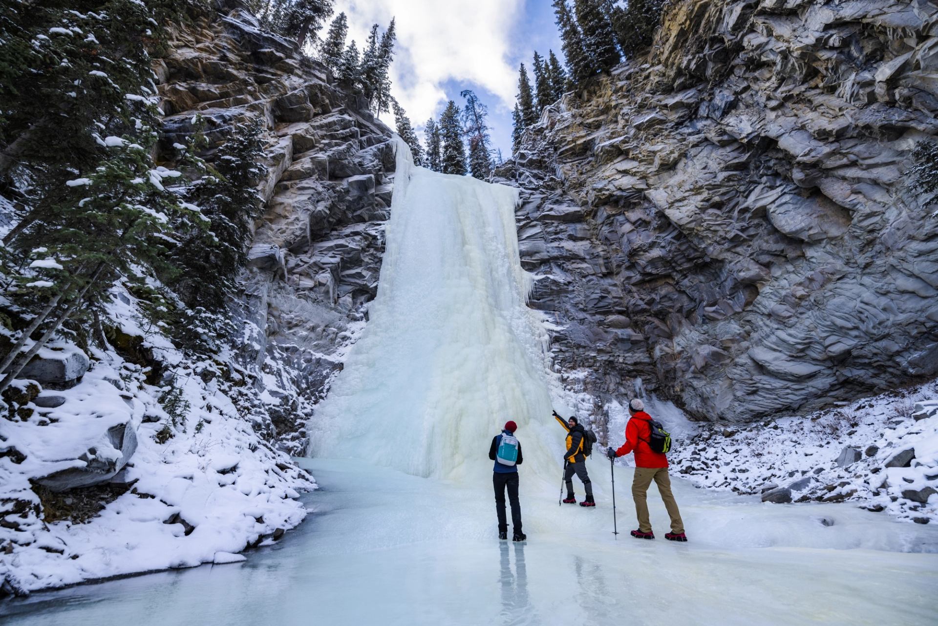 Three people stand on a frozen river below a towering ice waterfall, framed by rocky canyon walls and snow‑dusted trees.