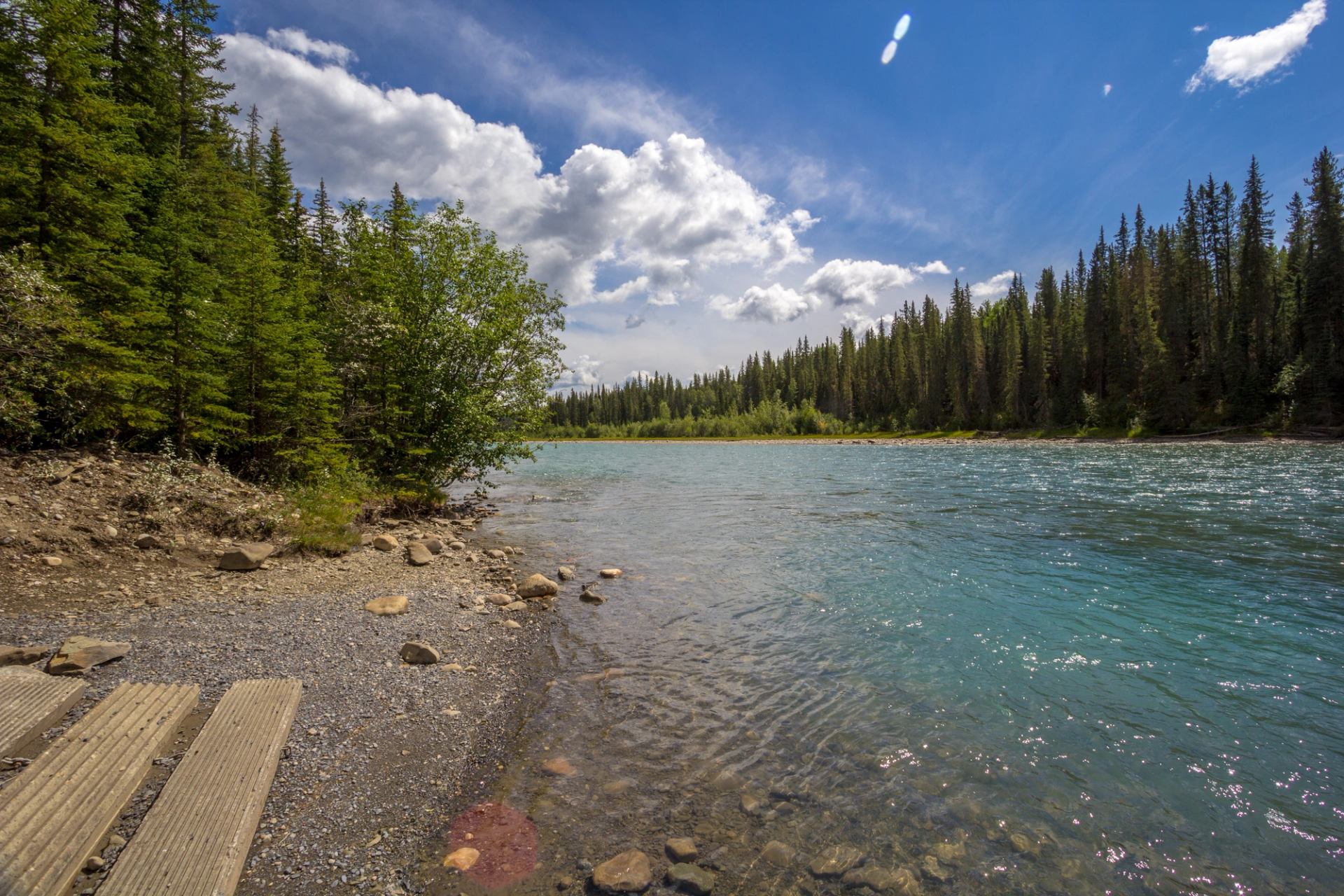 Rocky river shoreline with clear blue water, dense evergreen forest and cloudy sky.