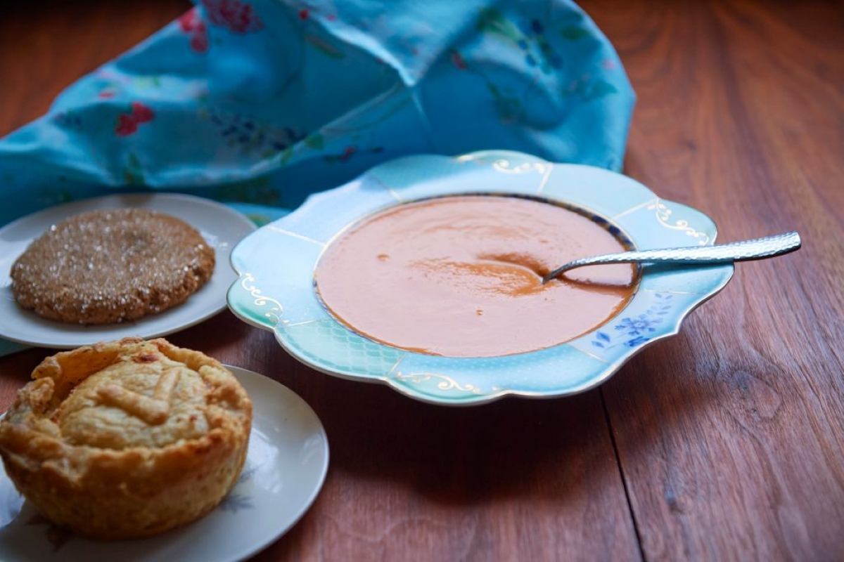 A light blue bowl of soup with a spoon, a small golden pie, and a sugared cookie on a wooden table with a blue patterned cloth.