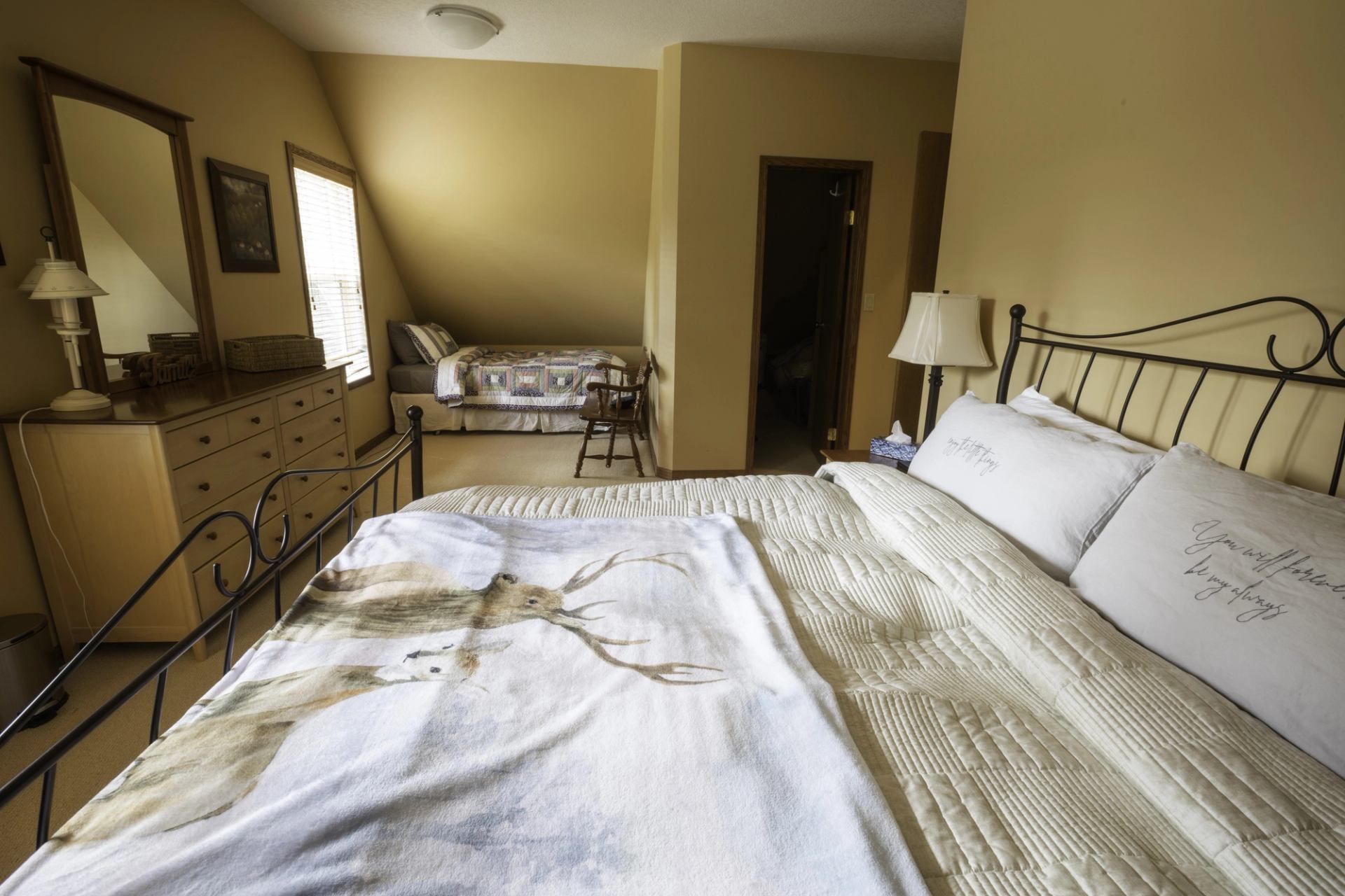 Cozy cabin bedroom with a king bed, dresser, and sitting area (twin bed) beneath a sloped ceiling