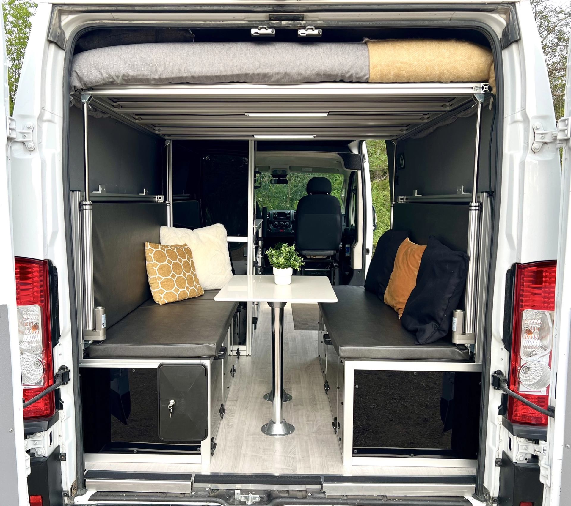 Camper van with benches, table, loft bed, and cozy interior view from rear doors.