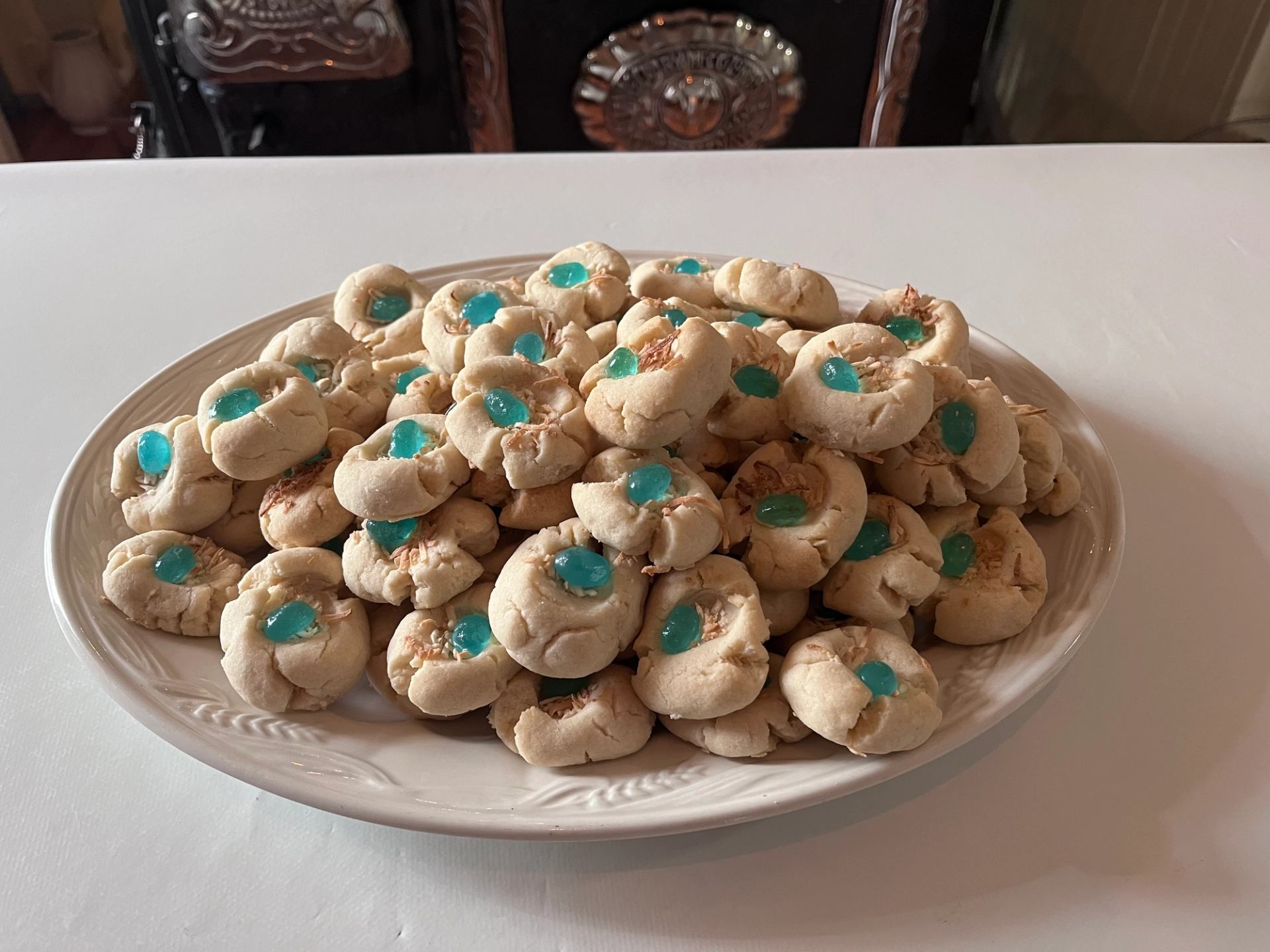A white plate filled with pale thumbprint-style cookies, each with a bright blue jelly center, and some topped with light brown flakes.
