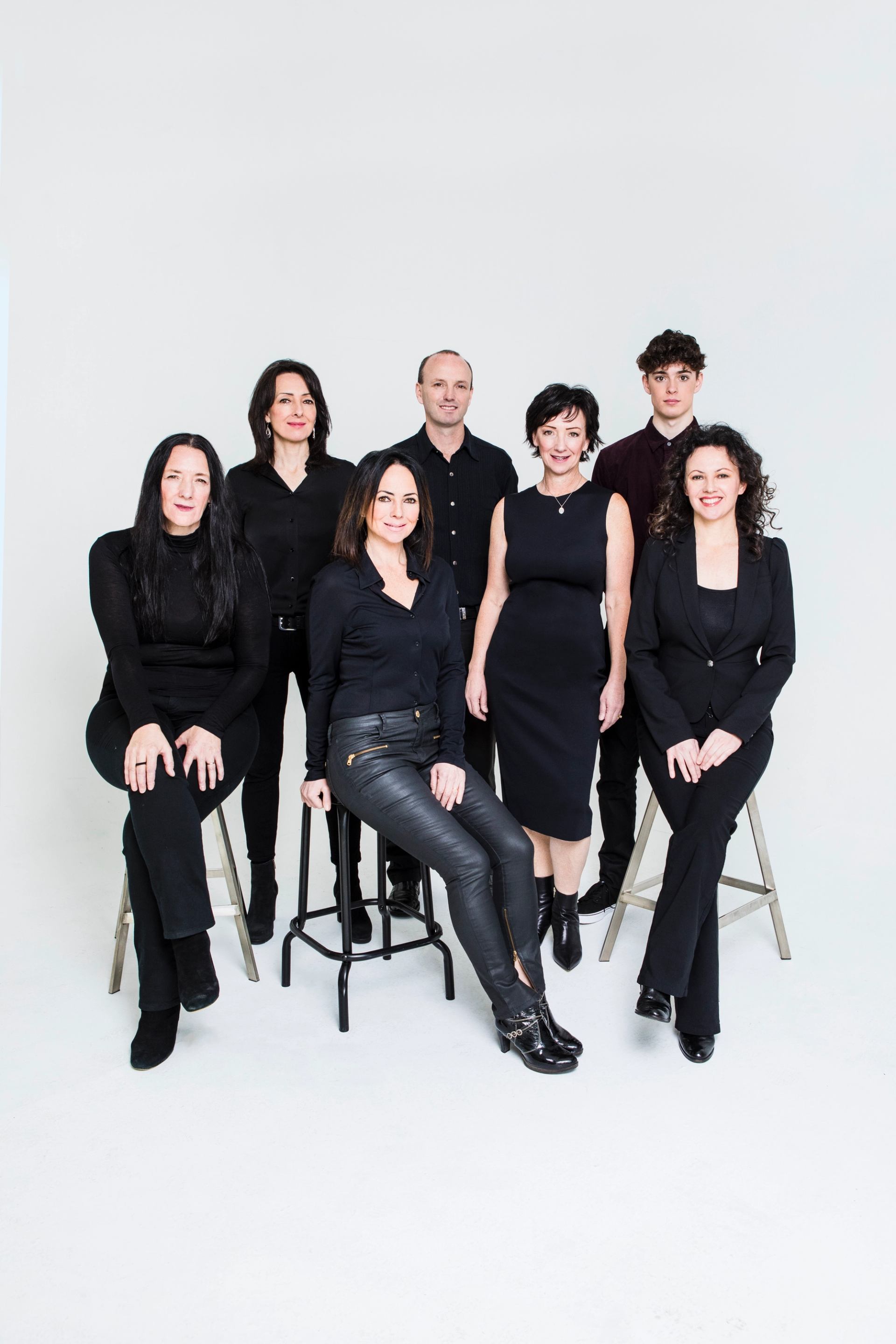 Group of seven people in black clothing posing together on stools against white background.
