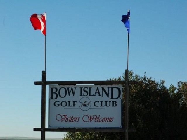 A sign for "Bow Island Golf Club, Visitors Welcome" with a Canadian and a blue flag flying above it under a clear blue sky.