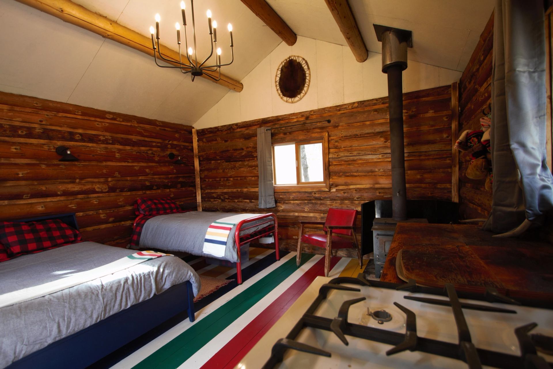 A rustic cabin bedroom with two beds, a wood stove, and colorful flooring.
