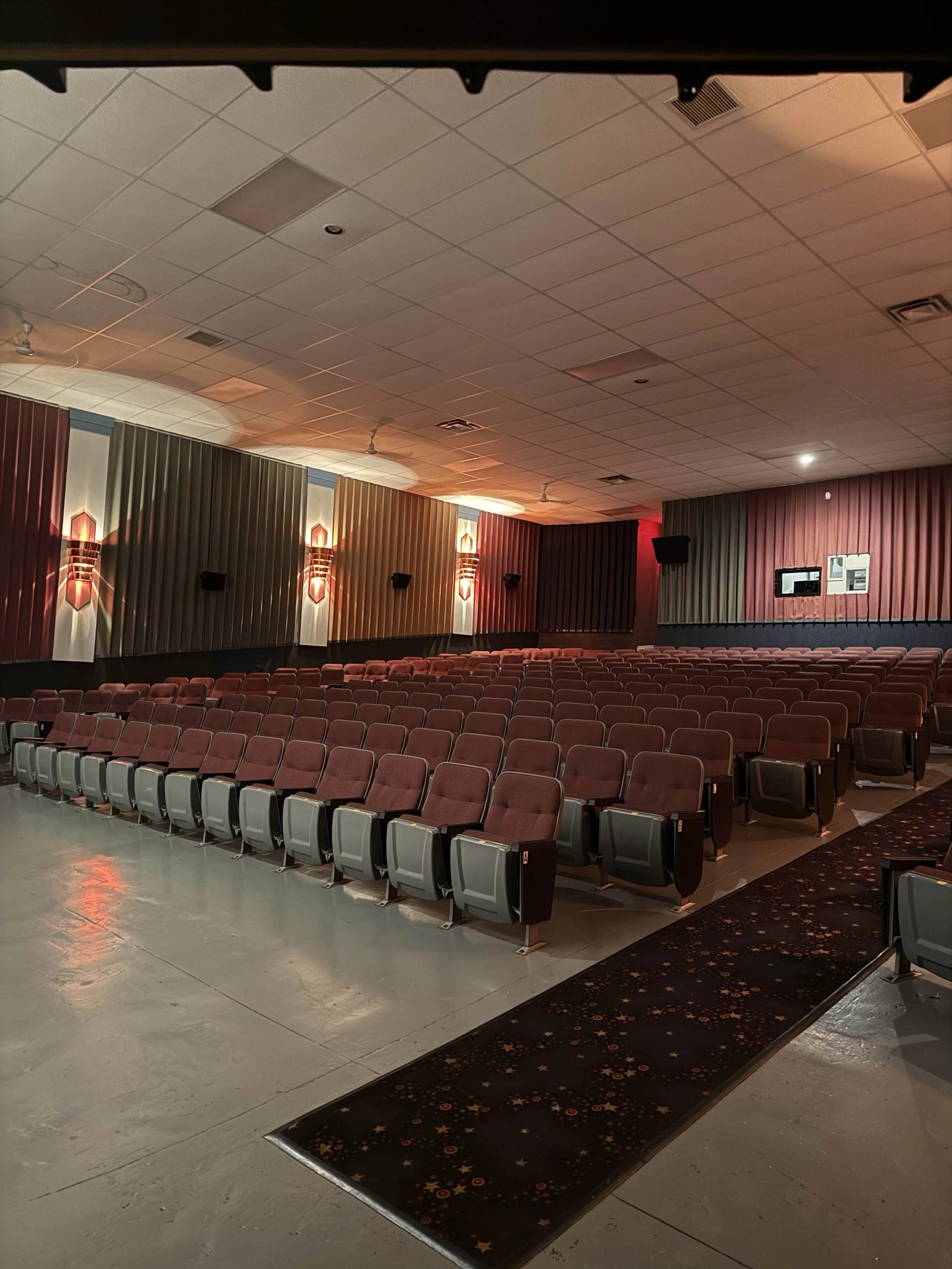 Cinema auditorium with rows of red seats, dim wall lights, and a central aisle.