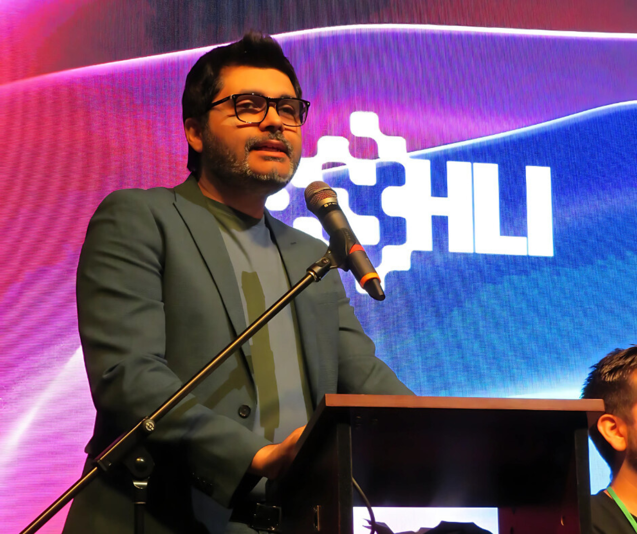 Person speaking at a podium on stage with a colorful digital backdrop.