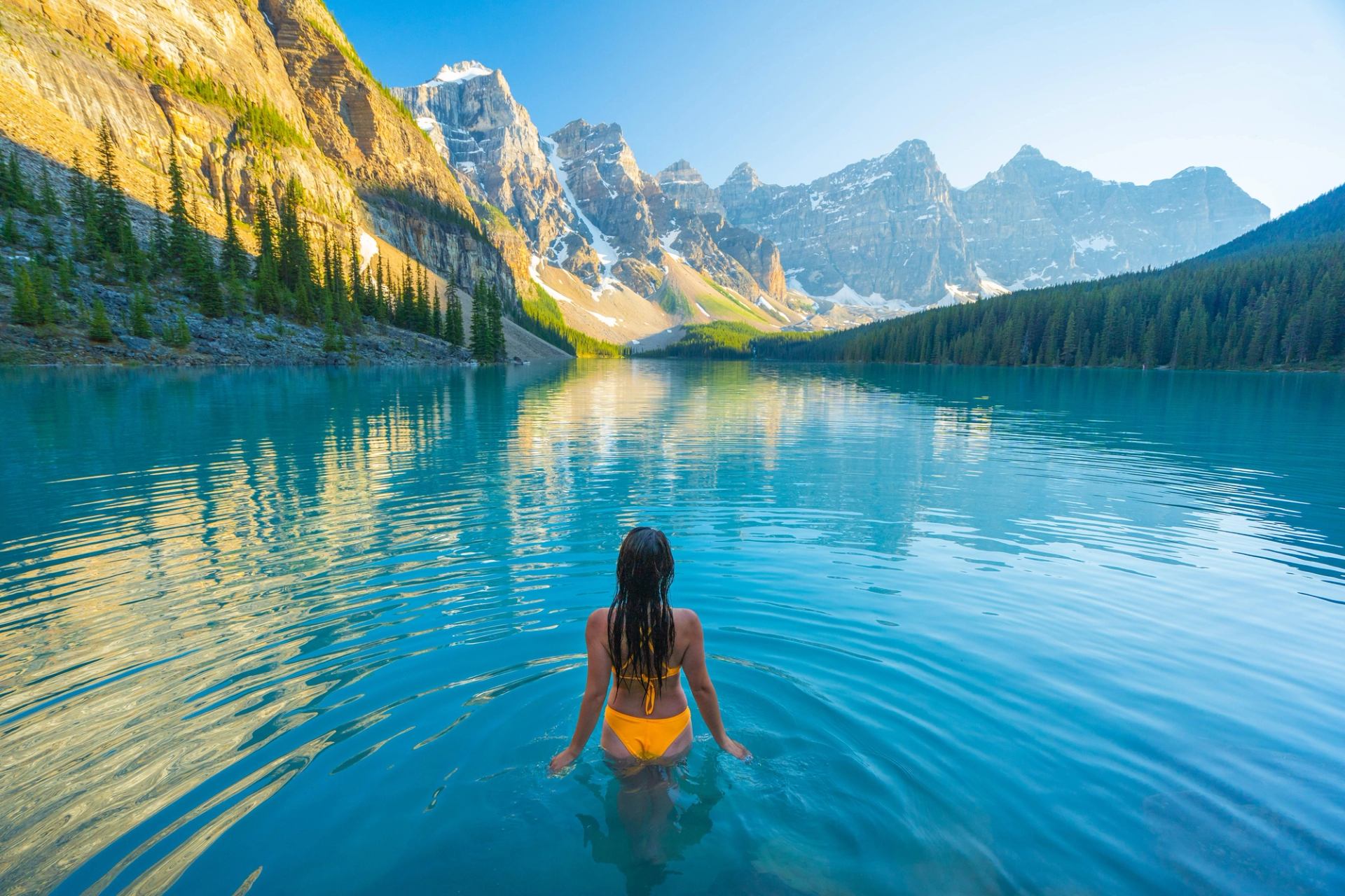 Person standing in Moraine Lake with mountain reflections and clear blue water.