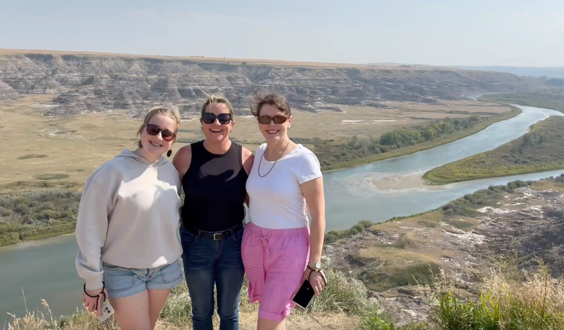 Three visitors stand at a scenic overlook above a winding river and dramatic badlands landscape.