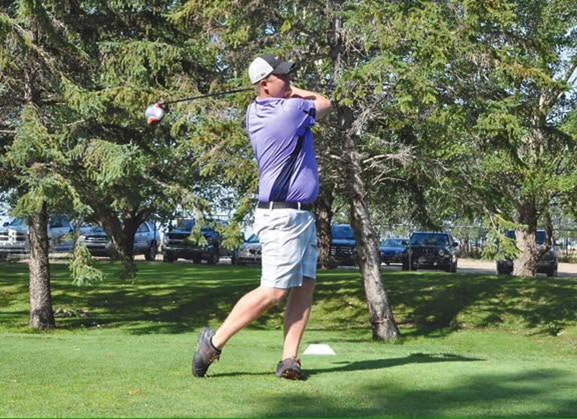 Golfer in purple shirt swings driver on tee box near trees and parked cars.