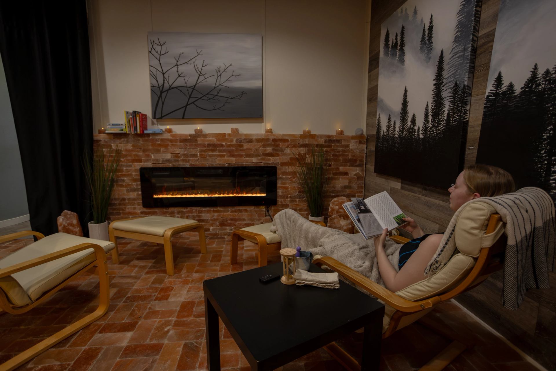 A woman relaxes in a lounge chair with her feet up, reading a book inside a cozy salt room. An elegant, modern electric fireplace glows against a brick wall, enhancing the peaceful ambiance.
