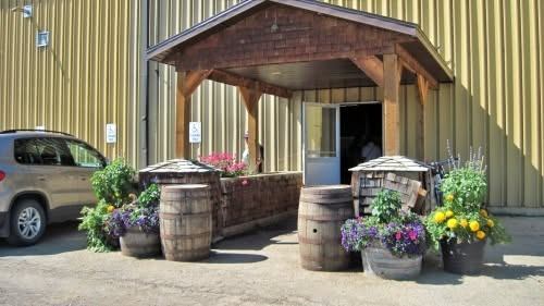 Olds Farmers' Market entrance features a covered doorway framed by flower planters and rustic barrels.