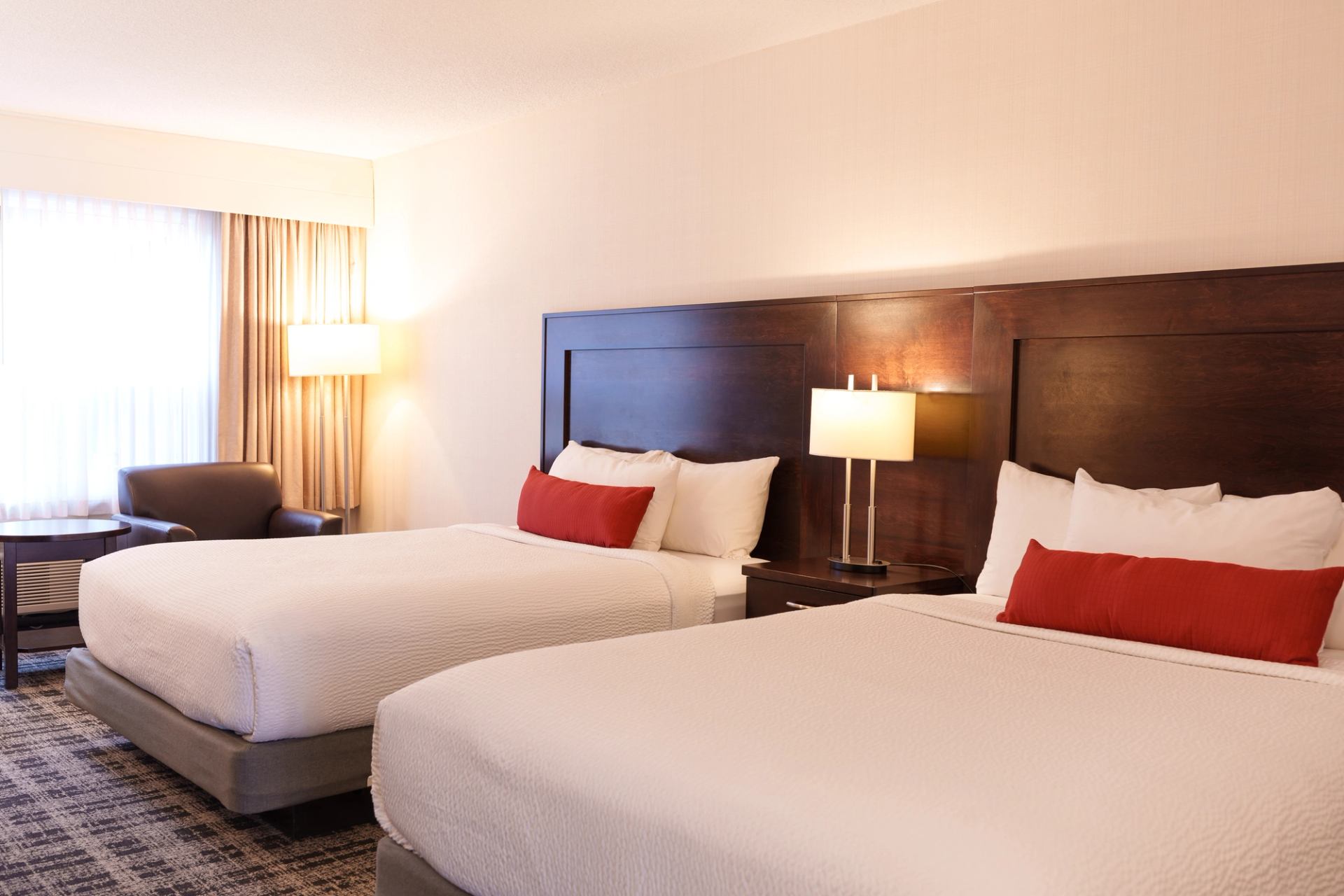 Hotel room with two beds, red accent pillows, bedside lamps and armchair by window.