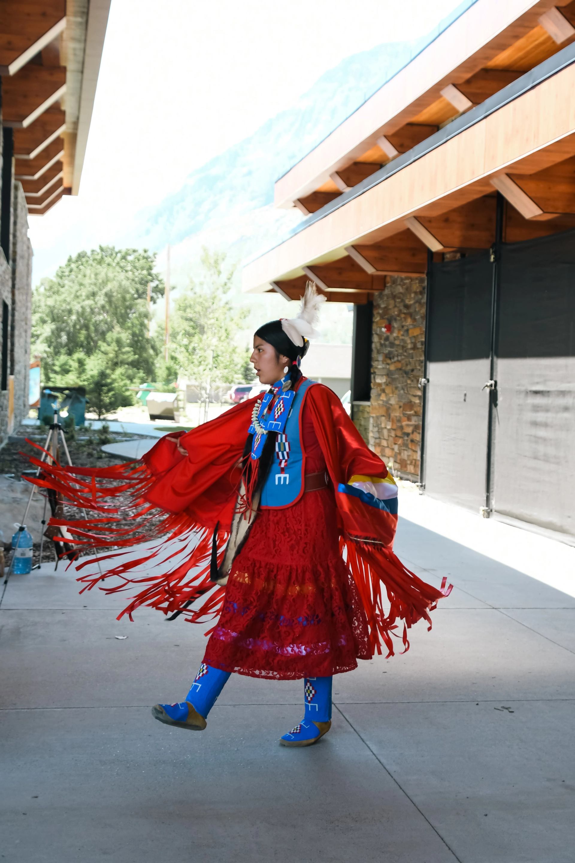 An Indigenous dancer in vibrant red regalia with fringe and beadwork performs outdoors. She wears a feather in her hair and blue beaded leggings, with buildings and trees in the background.
