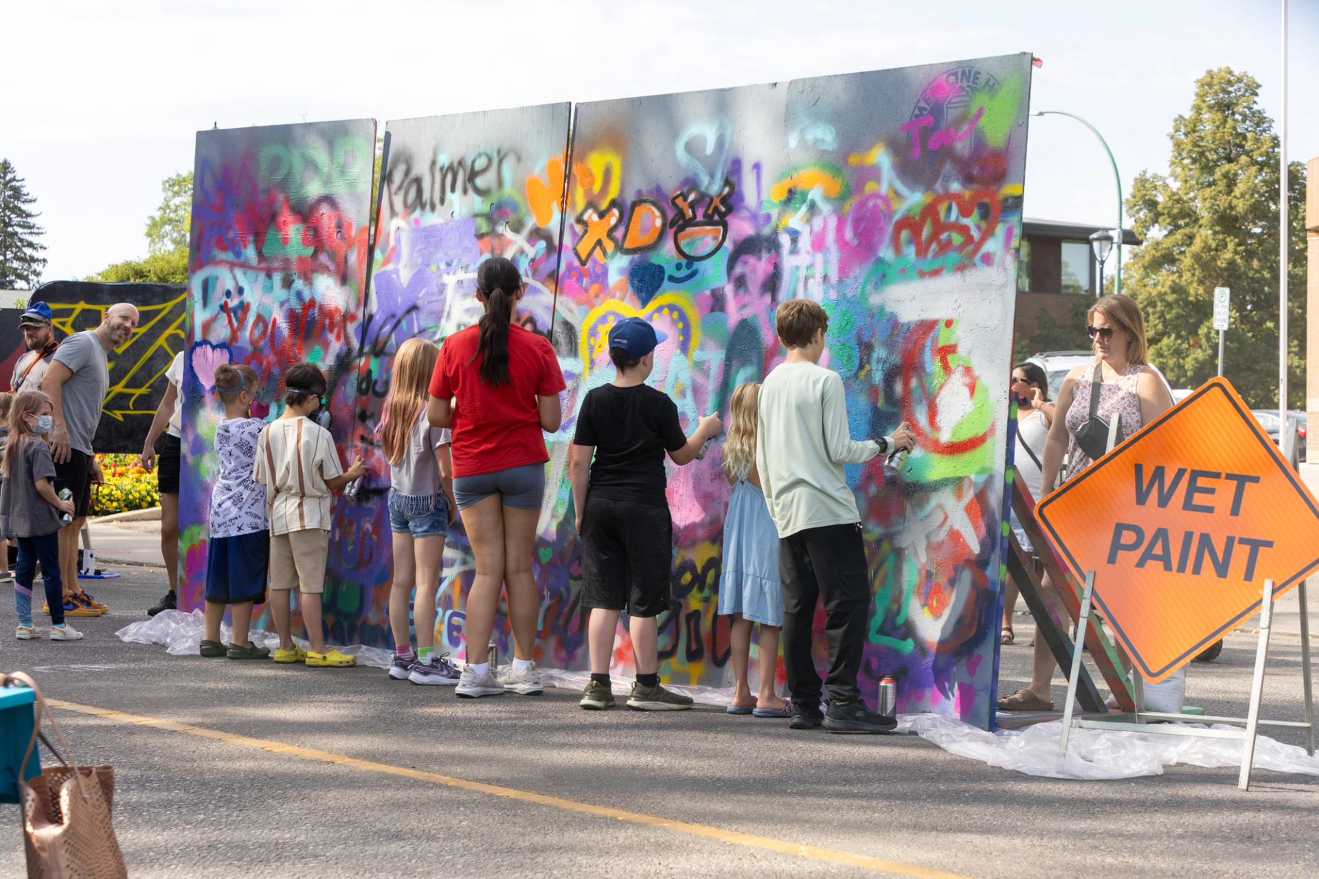 Children spray-paint a colourful graffiti wall at a street event beside wet paint sign.