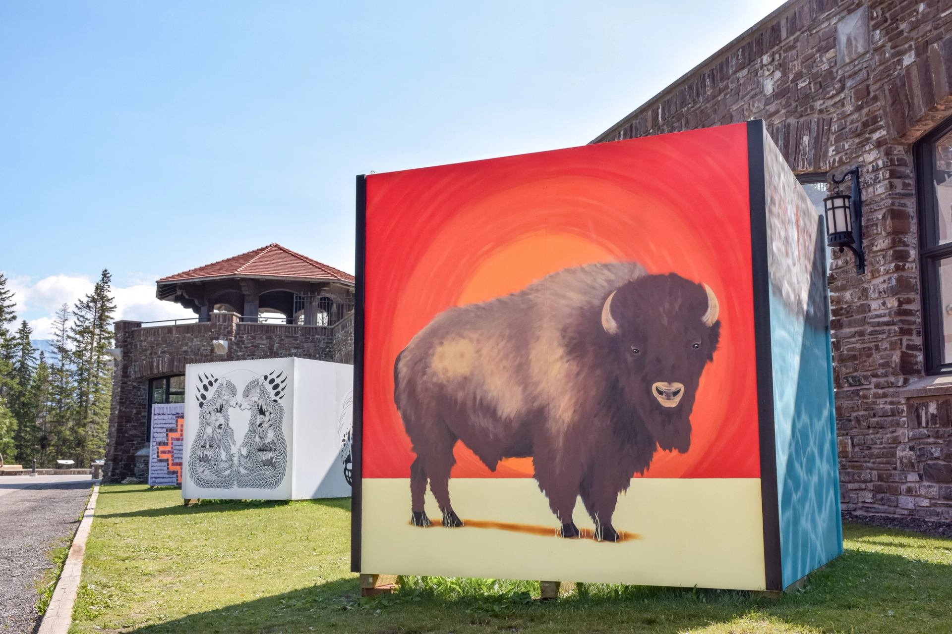 Large outdoor mural of a bison on red background beside stone building and trees.