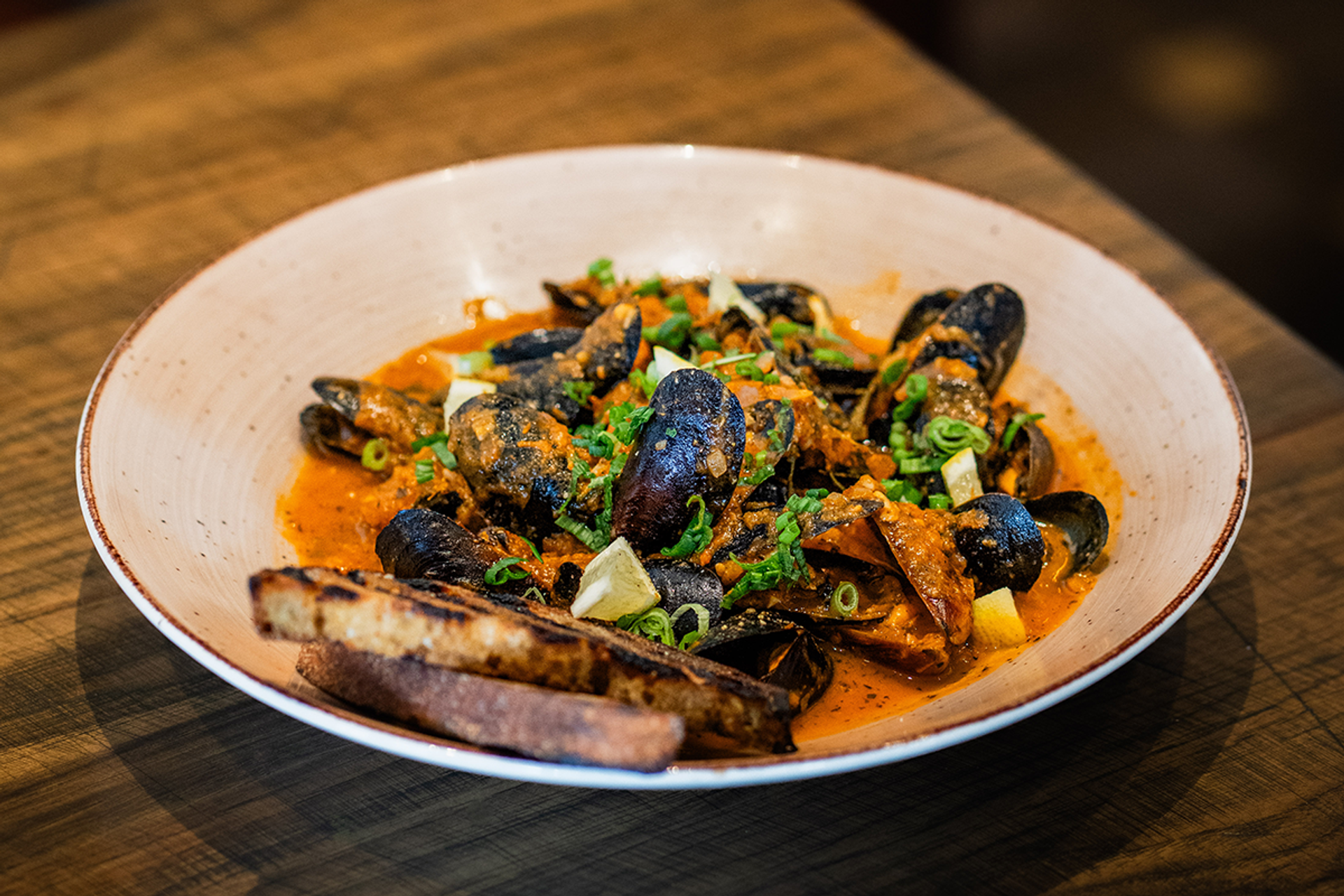 Bowl of mussels in orange sauce with chopped herbs and toasted bread slices.