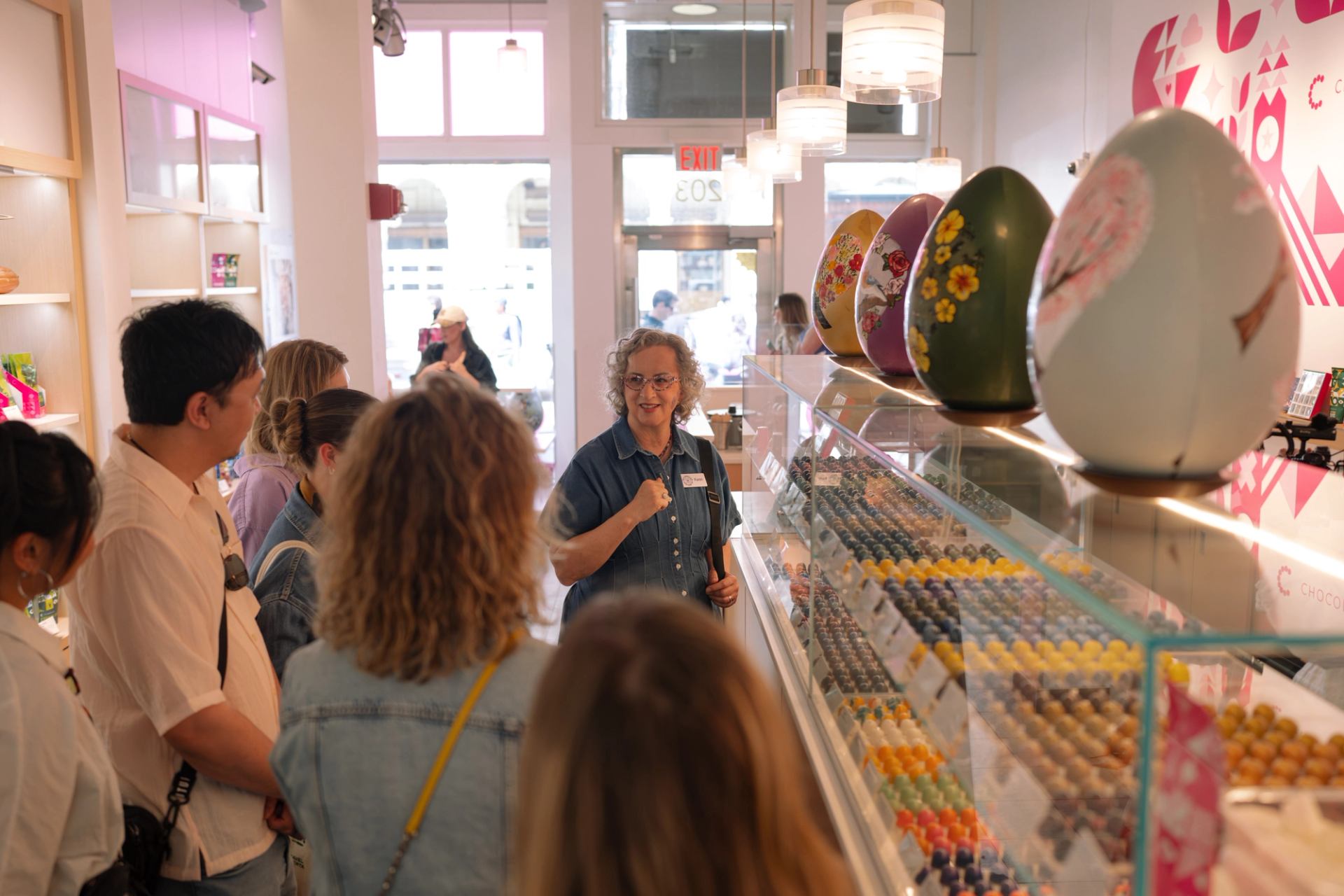 Chocolate shop staff member talks to visitors beside display of truffles and giant eggs.