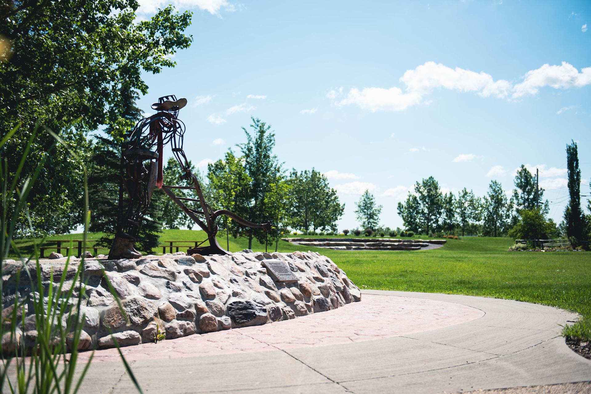Metal cowboy sculpture on stone base in grassy park with trees and blue sky.