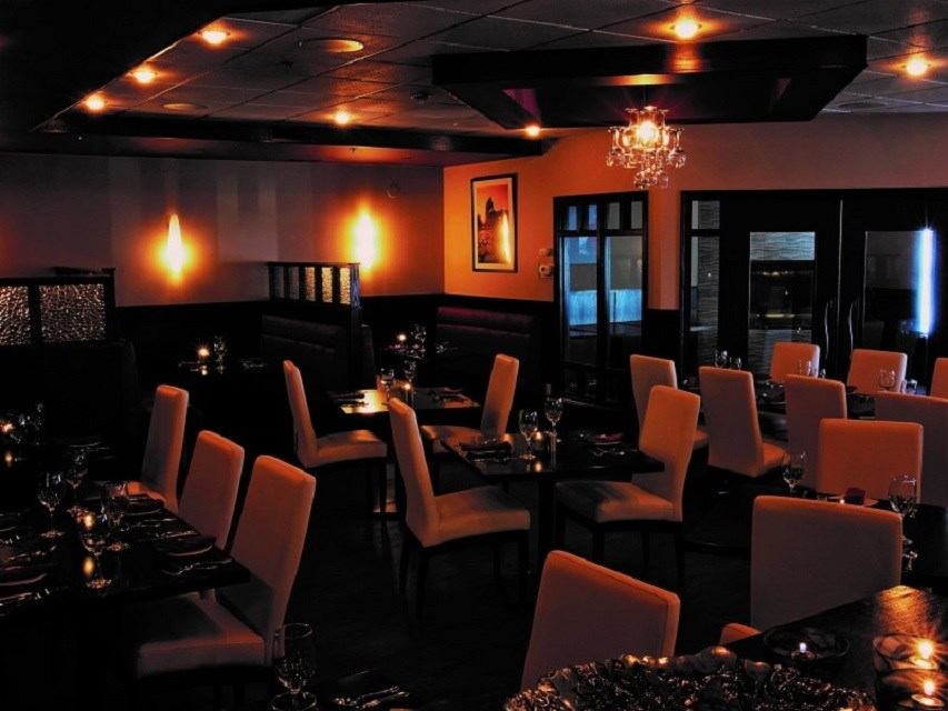 An empty, dimly lit restaurant dining room with warm orange walls, light-colored chairs, and set tables.