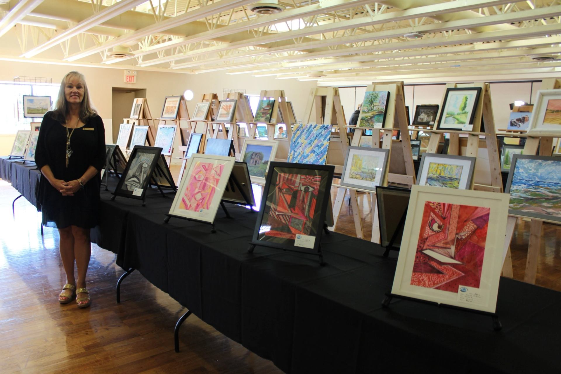 A smiling woman stands in an art exhibition with numerous paintings displayed on easels and tables.