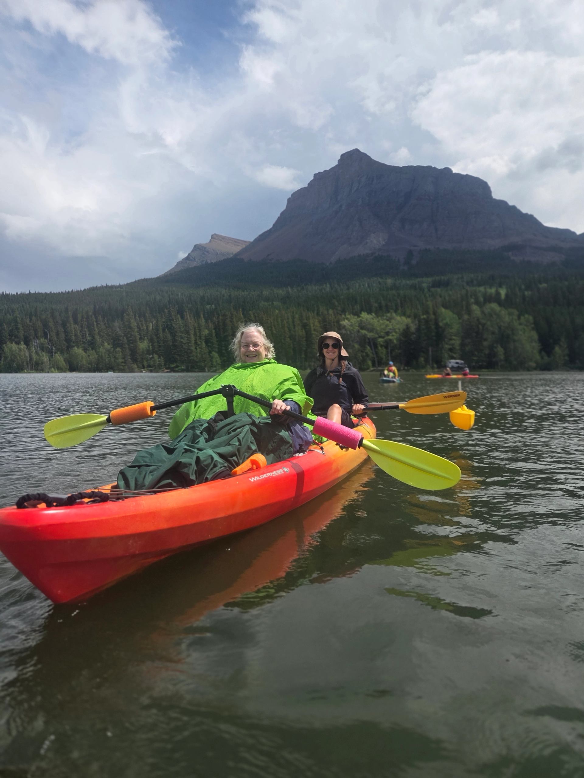 Two people kayaking on a calm lake with a mountain backdrop and forested shoreline.