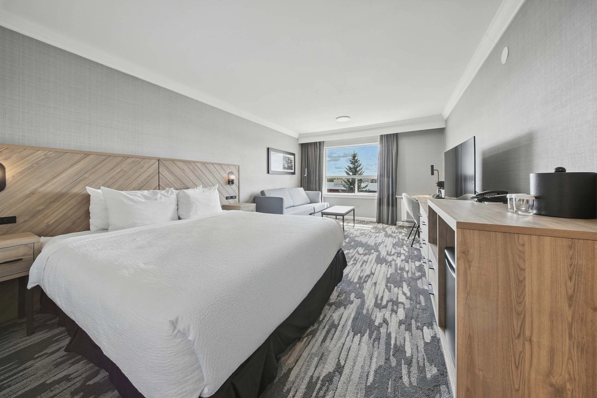 Hotel room with king bed, sofa, desk, television and window overlooking buildings.