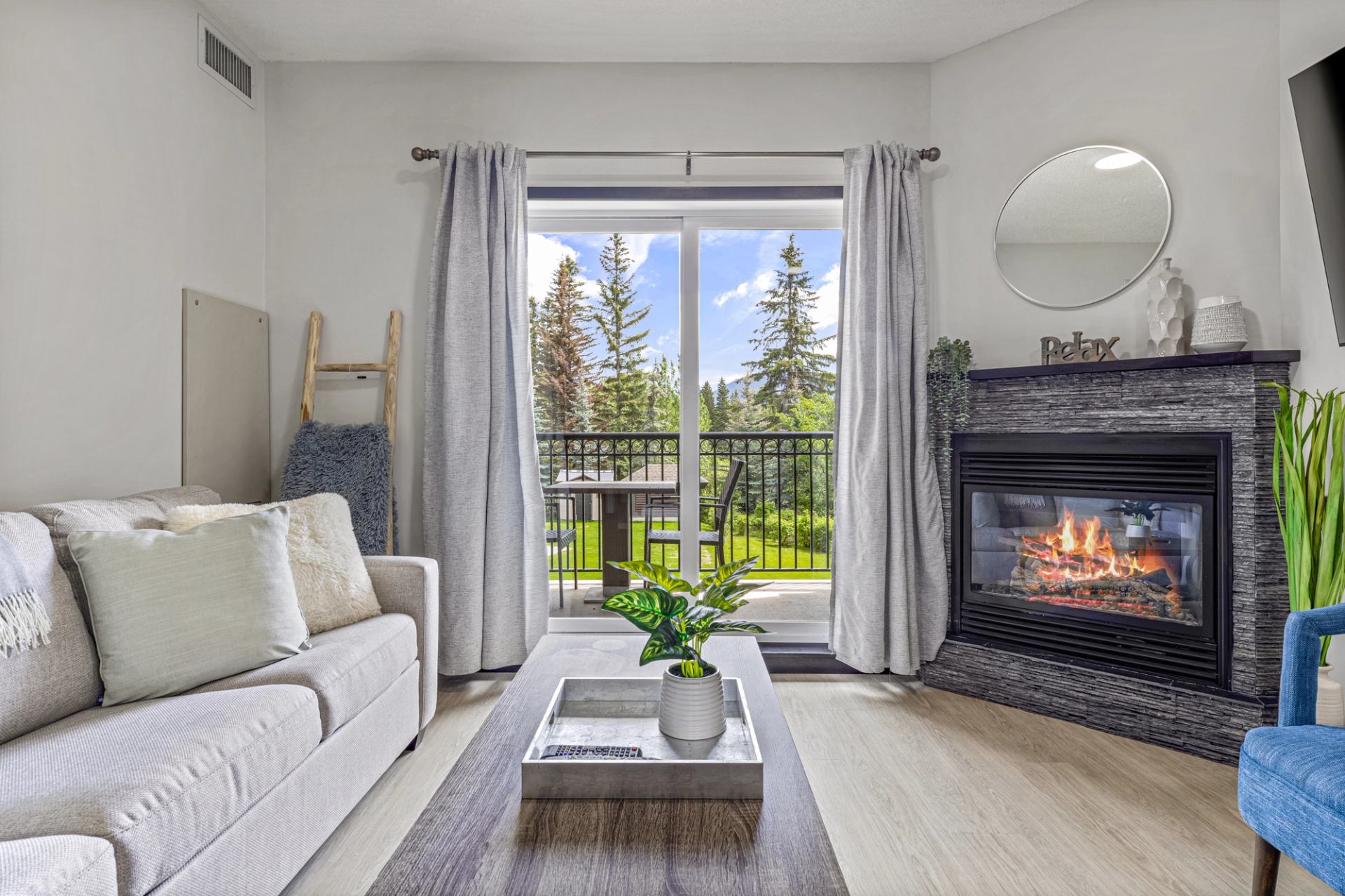 Living room with sofa, lit fireplace and balcony doors overlooking evergreen trees.