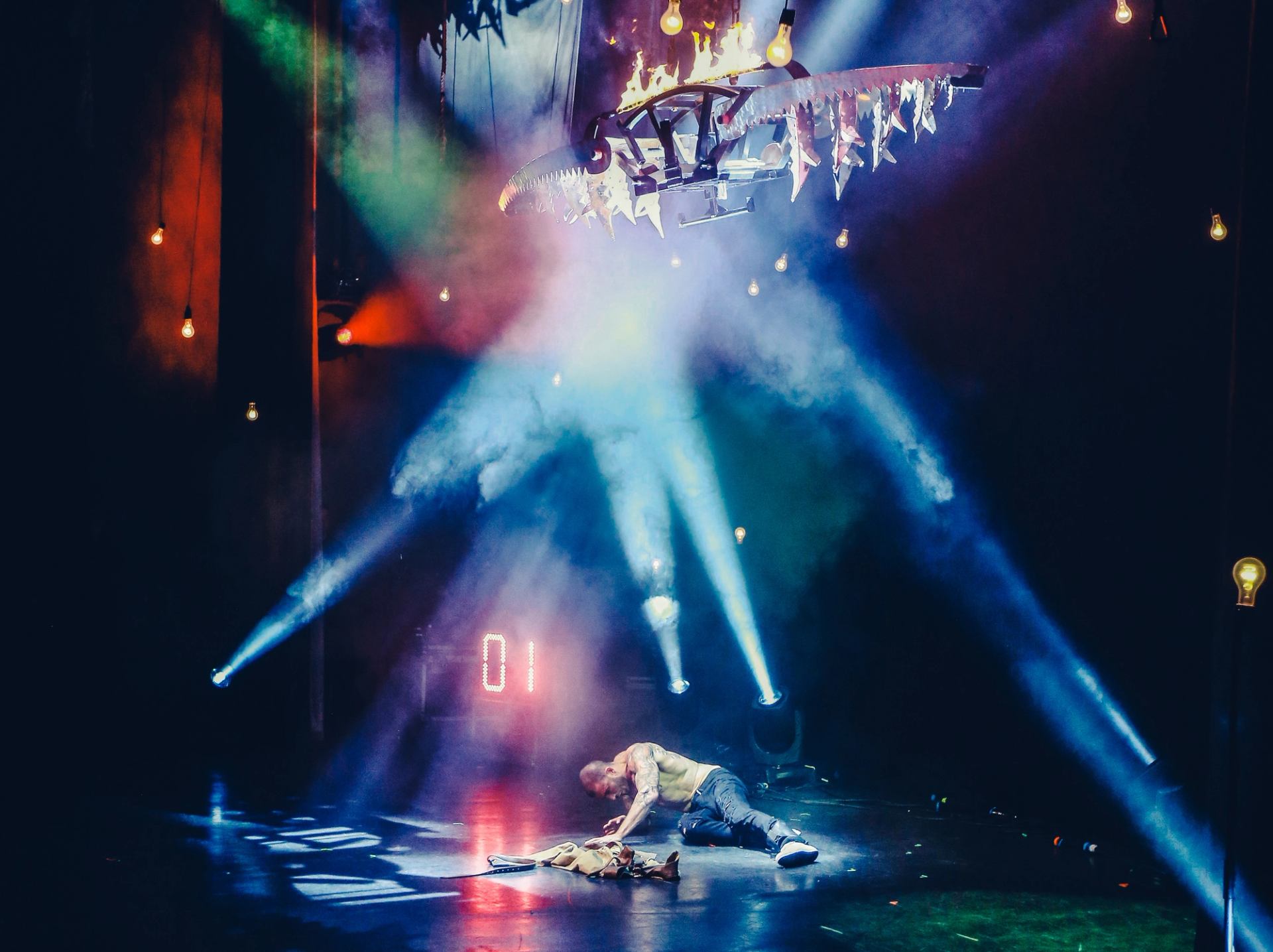Stage performer lies on floor under flaming circular rig and colourful spotlights.