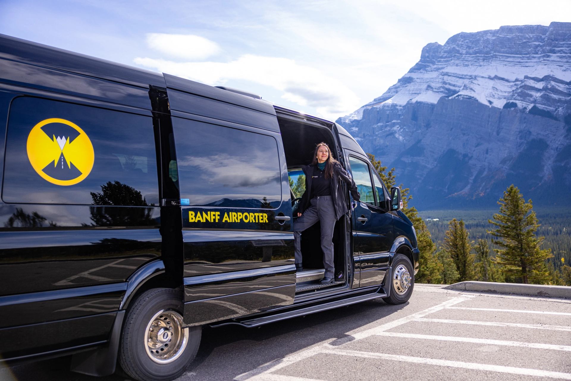 Driver stands in a Banff Airporter van doorway before a snow-covered mountain.