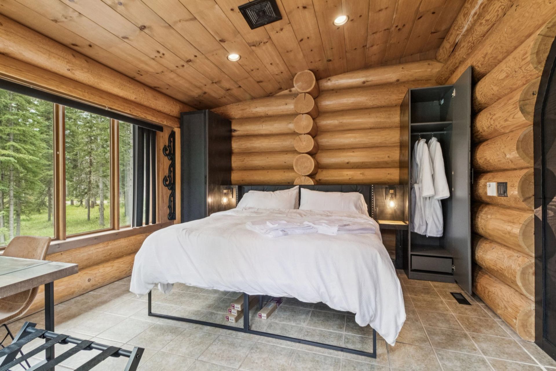 Log cabin bedroom with white bedding, robes, and forest views.