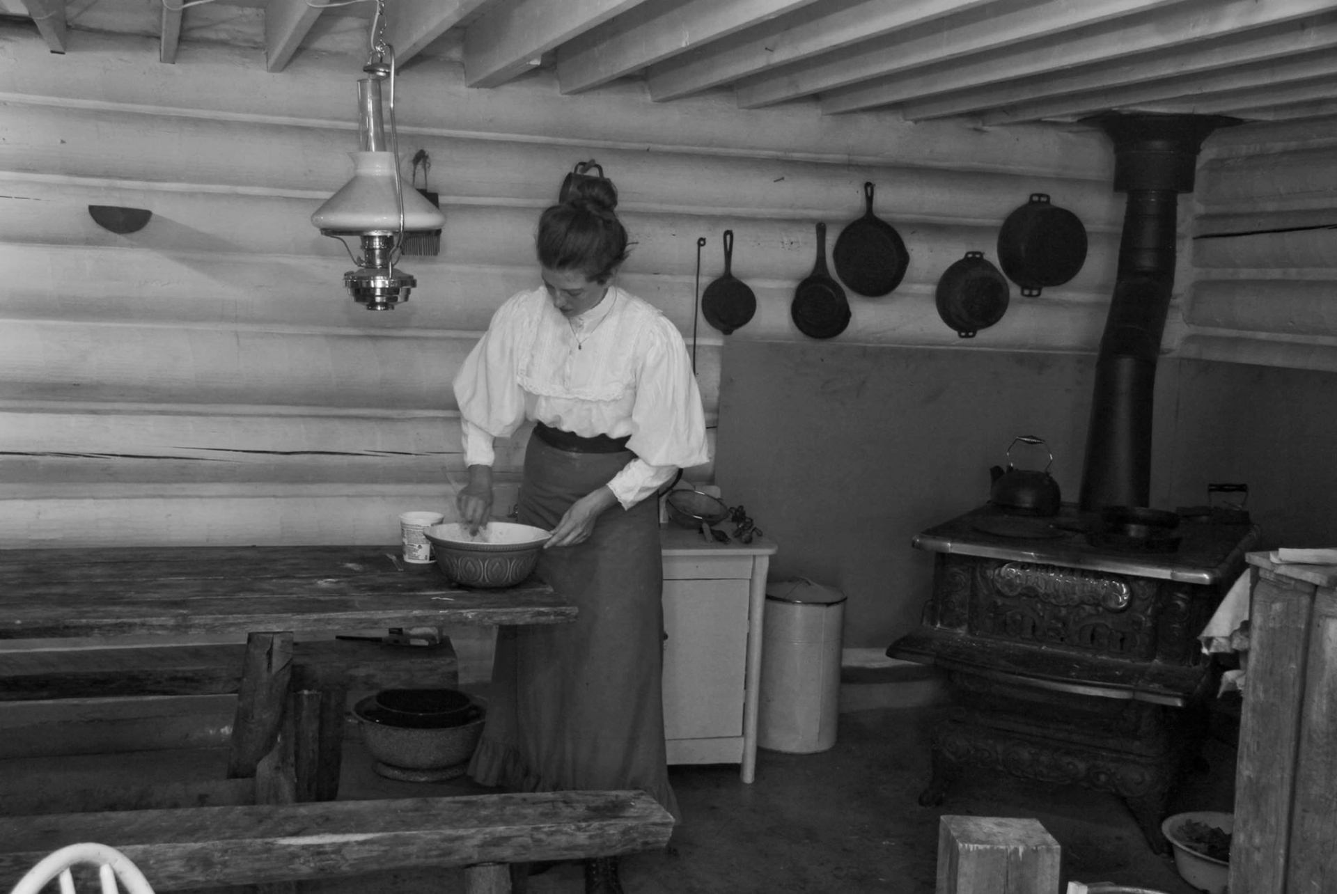 An Interpreter in a historical outfit stirs the contents of a bowl on the table in front of the wood stove.