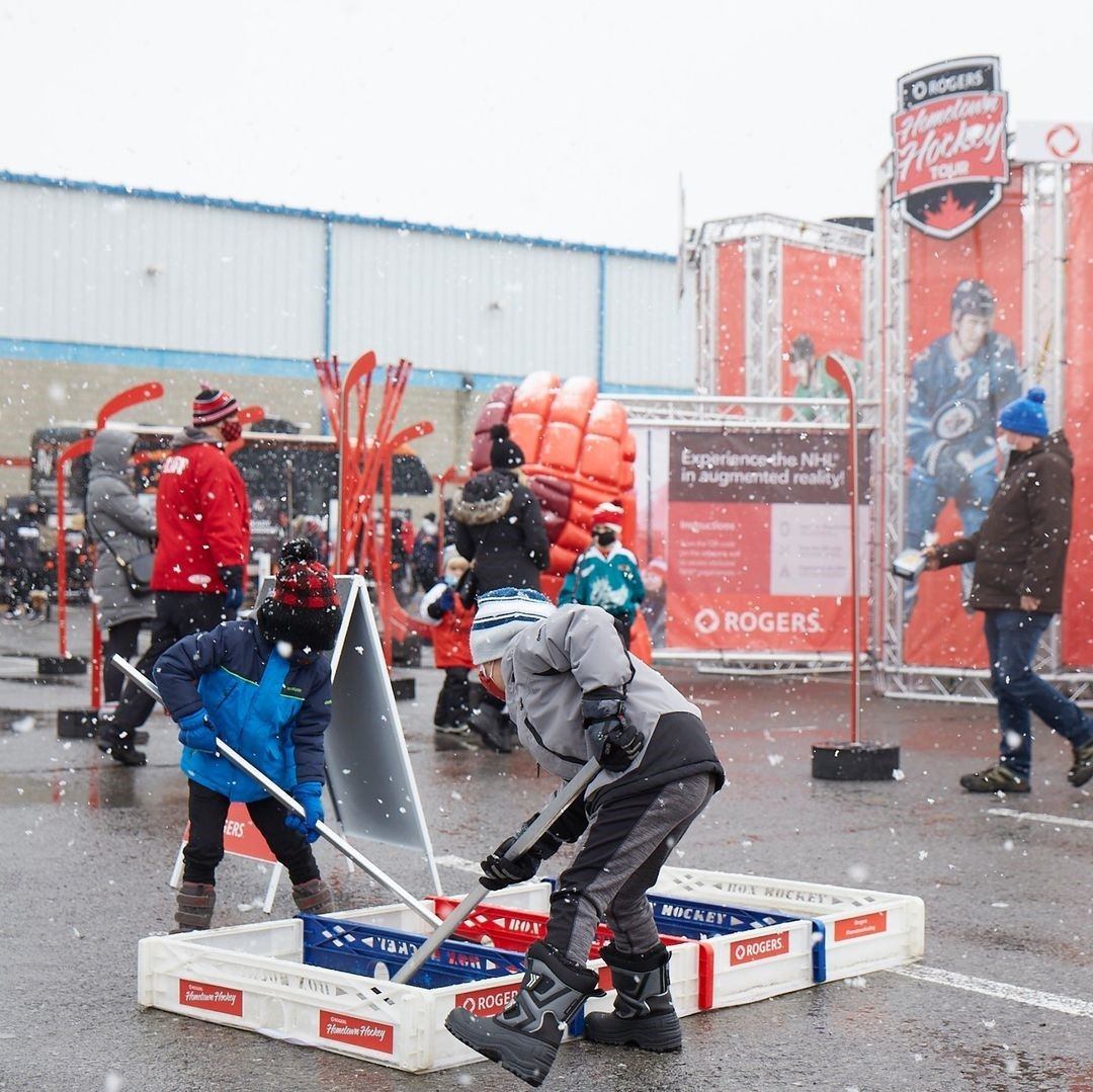 People play street hockey amid falling snow beside Rogers banners and inflatable glove.