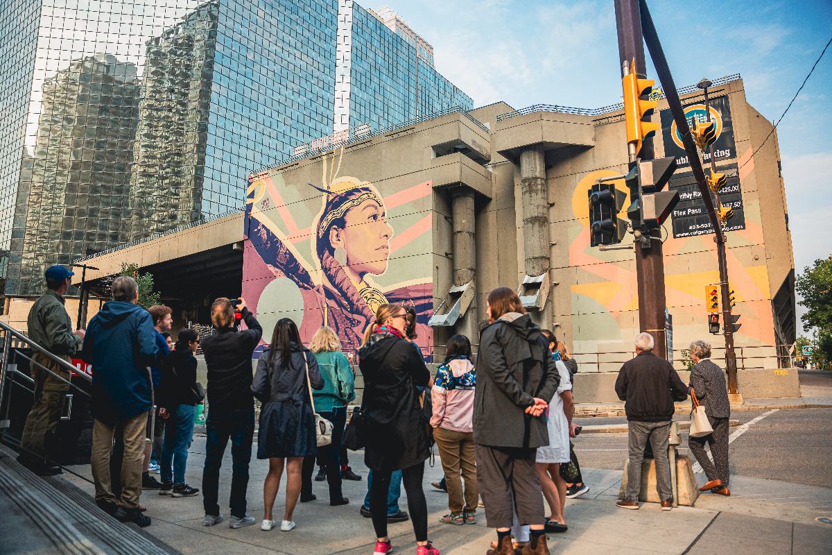 Small crowd gathers before pink and blue mural depicting a Cree woman in traditional regalia.