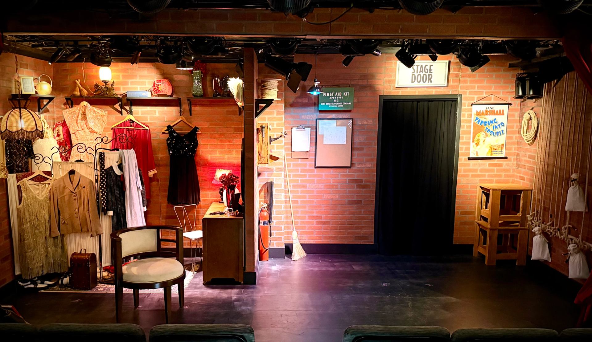 Theatre stage with vintage costumes, props and a black door against brick walls.