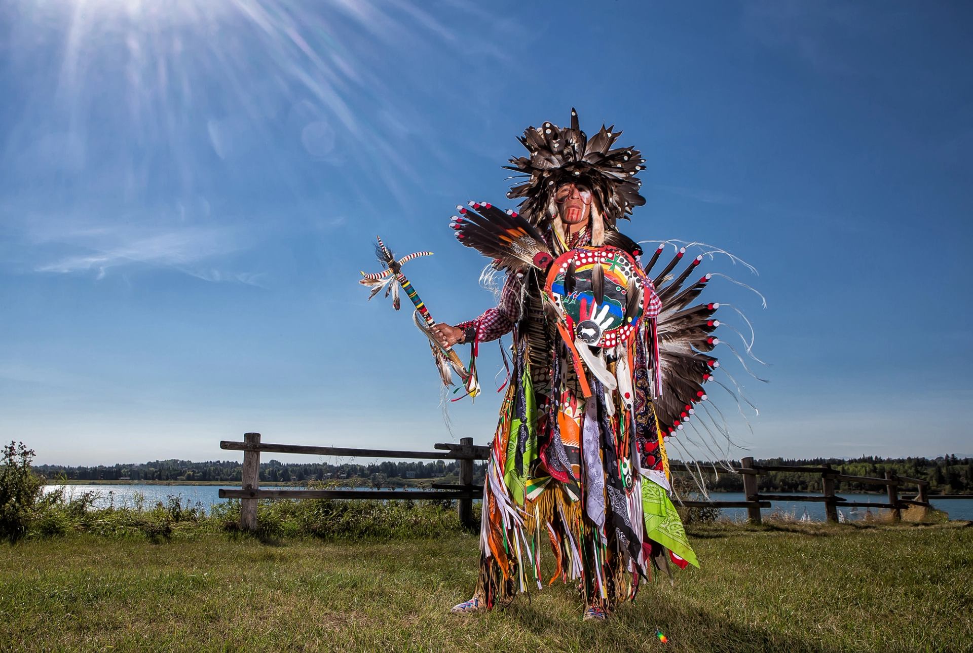 Indigenous dancer in colourful regalia stands by lakeside fence under bright blue sky.