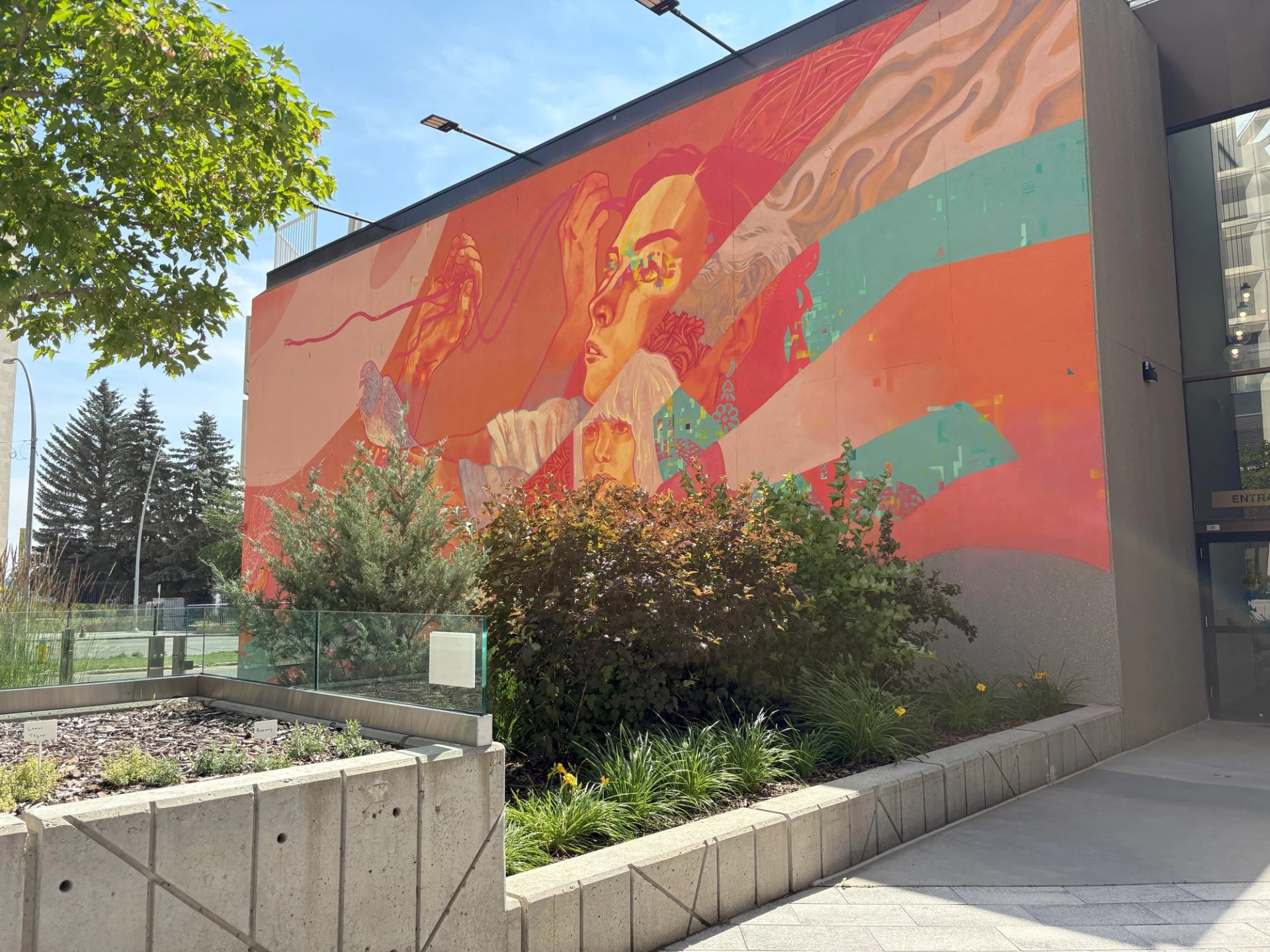 Colorful mural with abstract shapes and figures on building wall, plants in foreground.