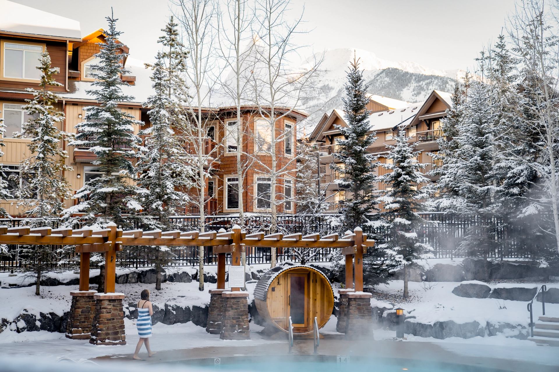 Snow-covered lodge buildings and evergreens surround a steaming outdoor pool and barrel sauna.
