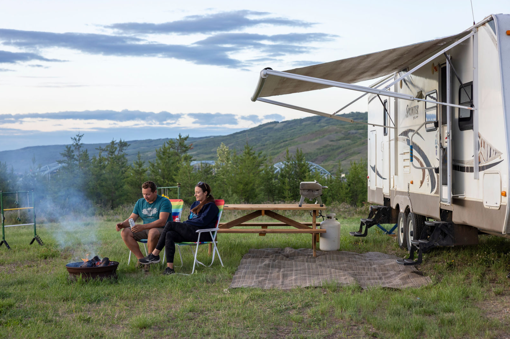 Campers sit by a fire beside an RV and picnic table at a grassy campground with hills in the background.