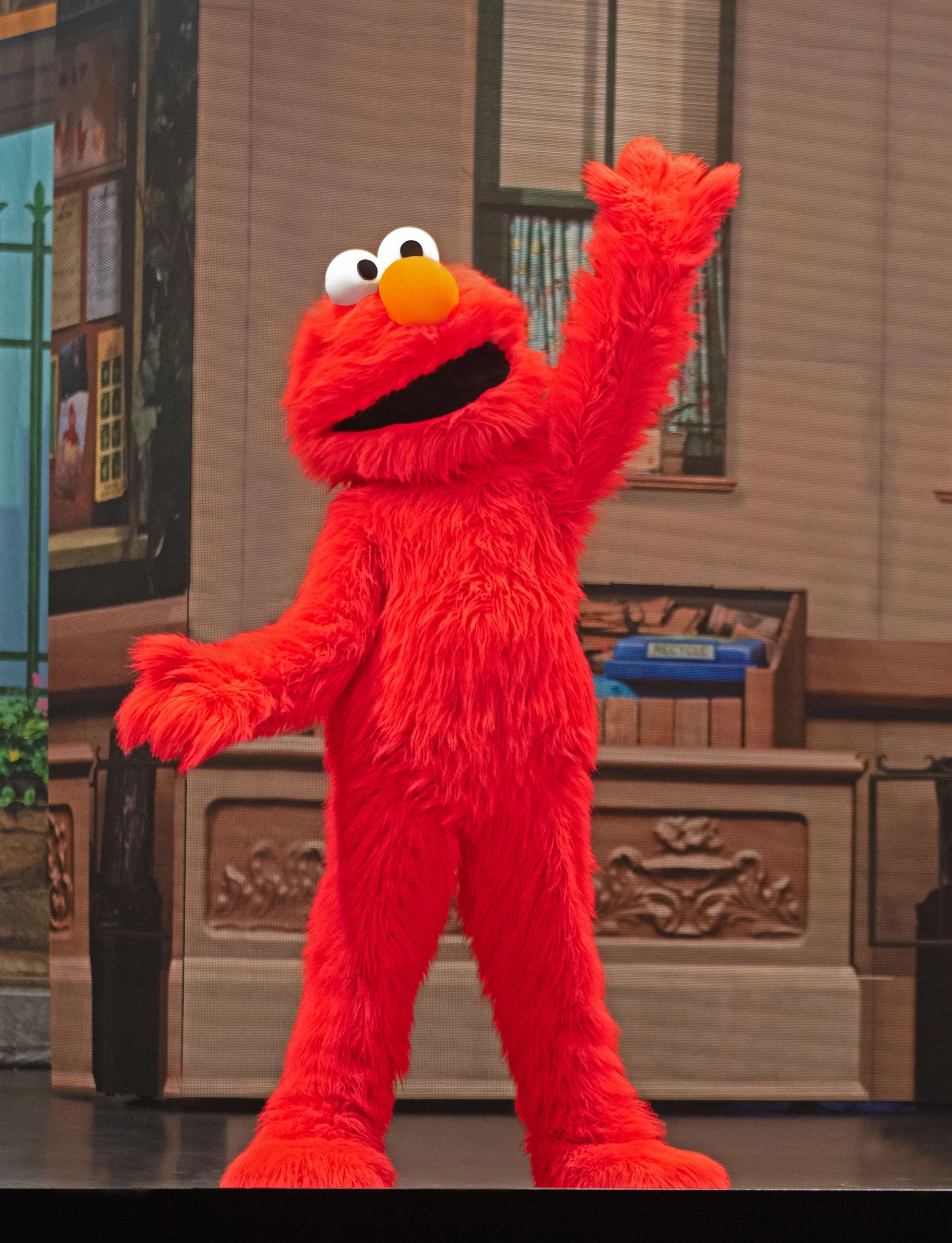 Red furry puppet character Elmo stands on stage, raising one arm in greeting.