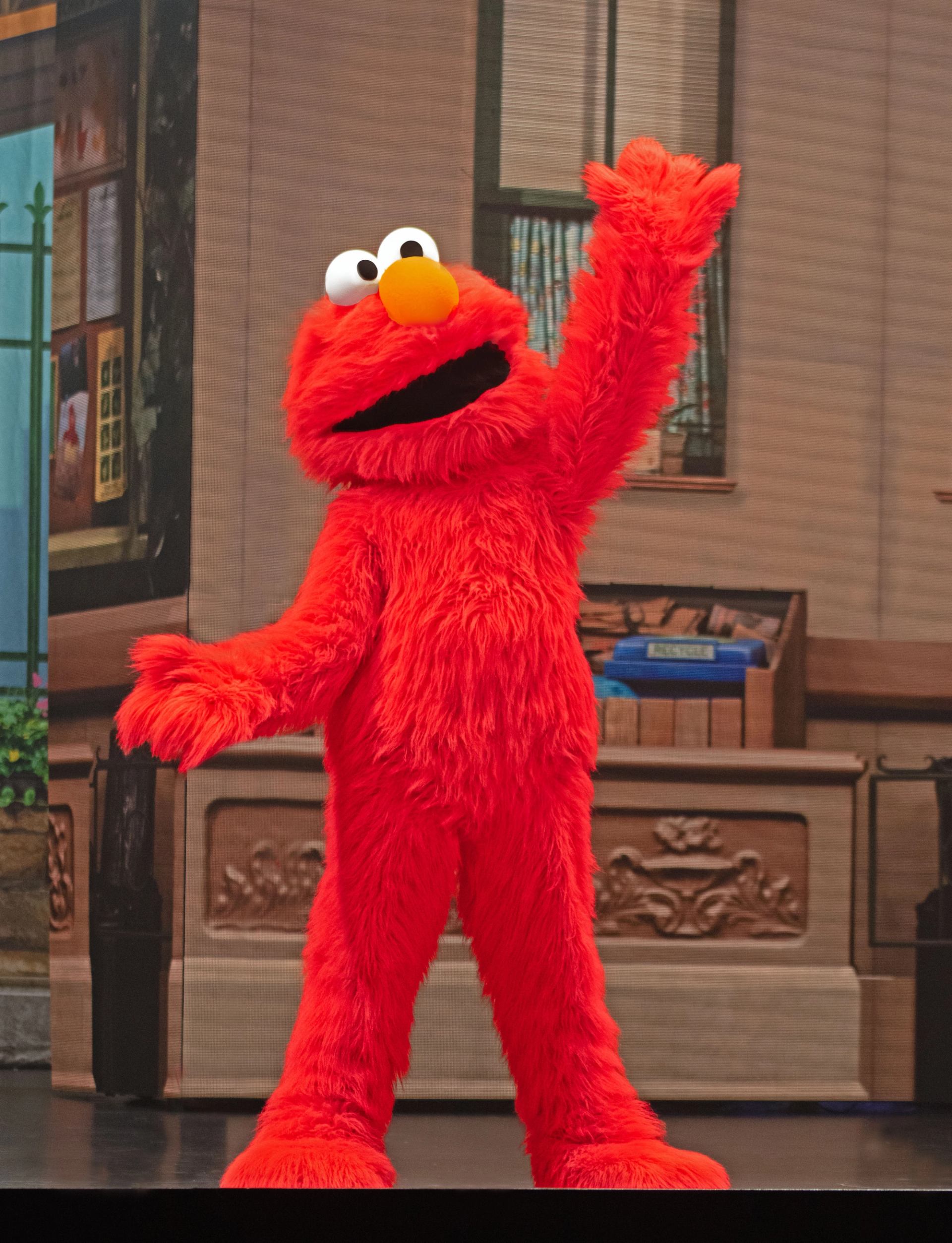 Red furry puppet character Elmo stands on stage, raising one arm in greeting.