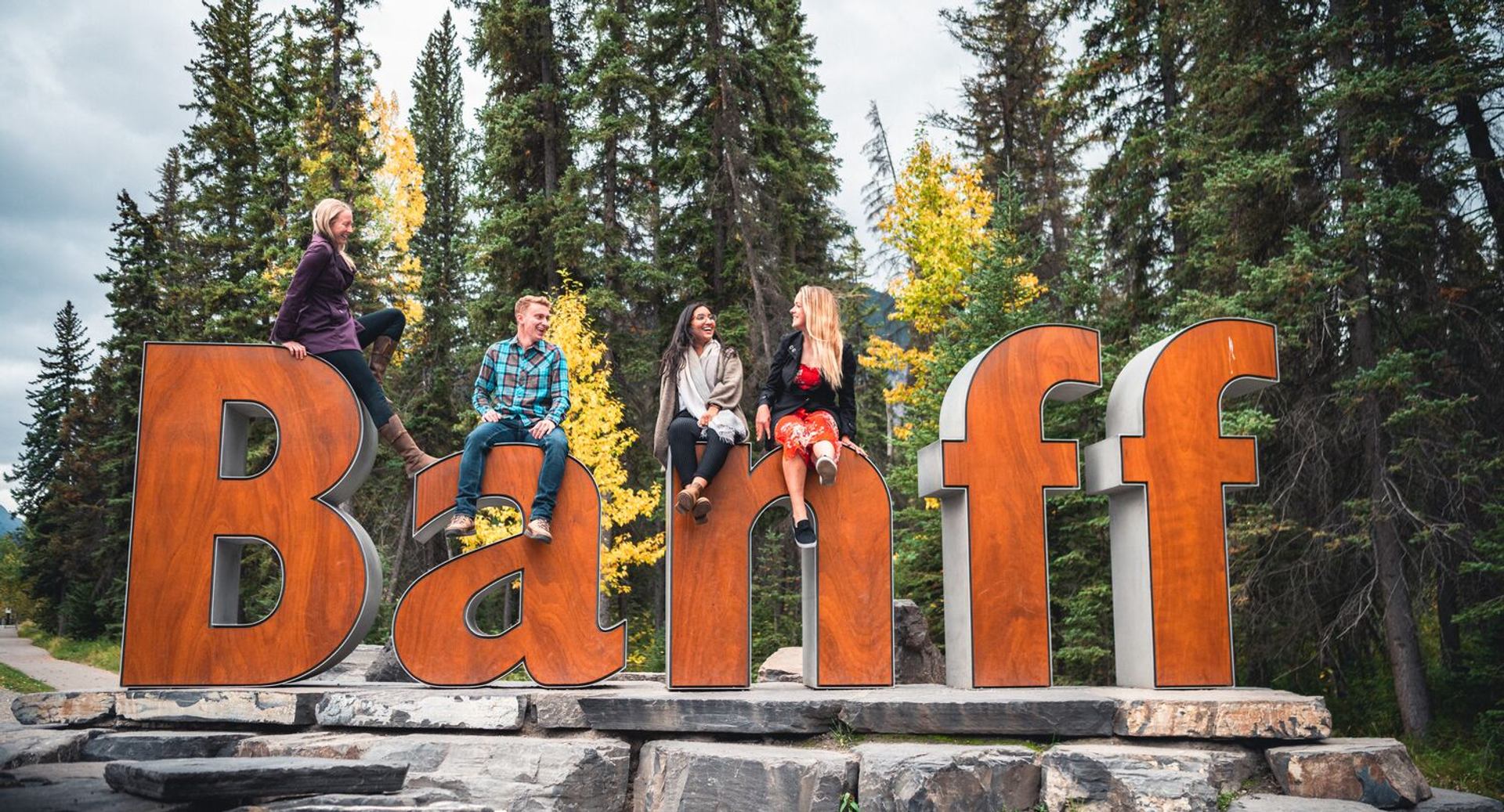 Large orange Banff sign with people sitting on letters, surrounded by forest trees.