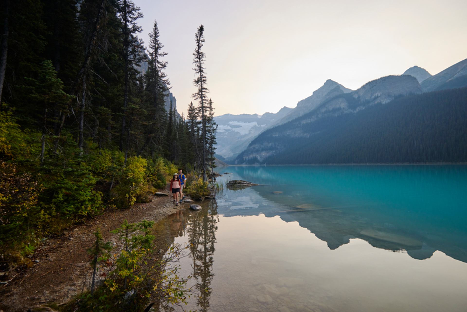 Two visitors walk along a forested lakeshore with turquoise water and mountains in the distance.