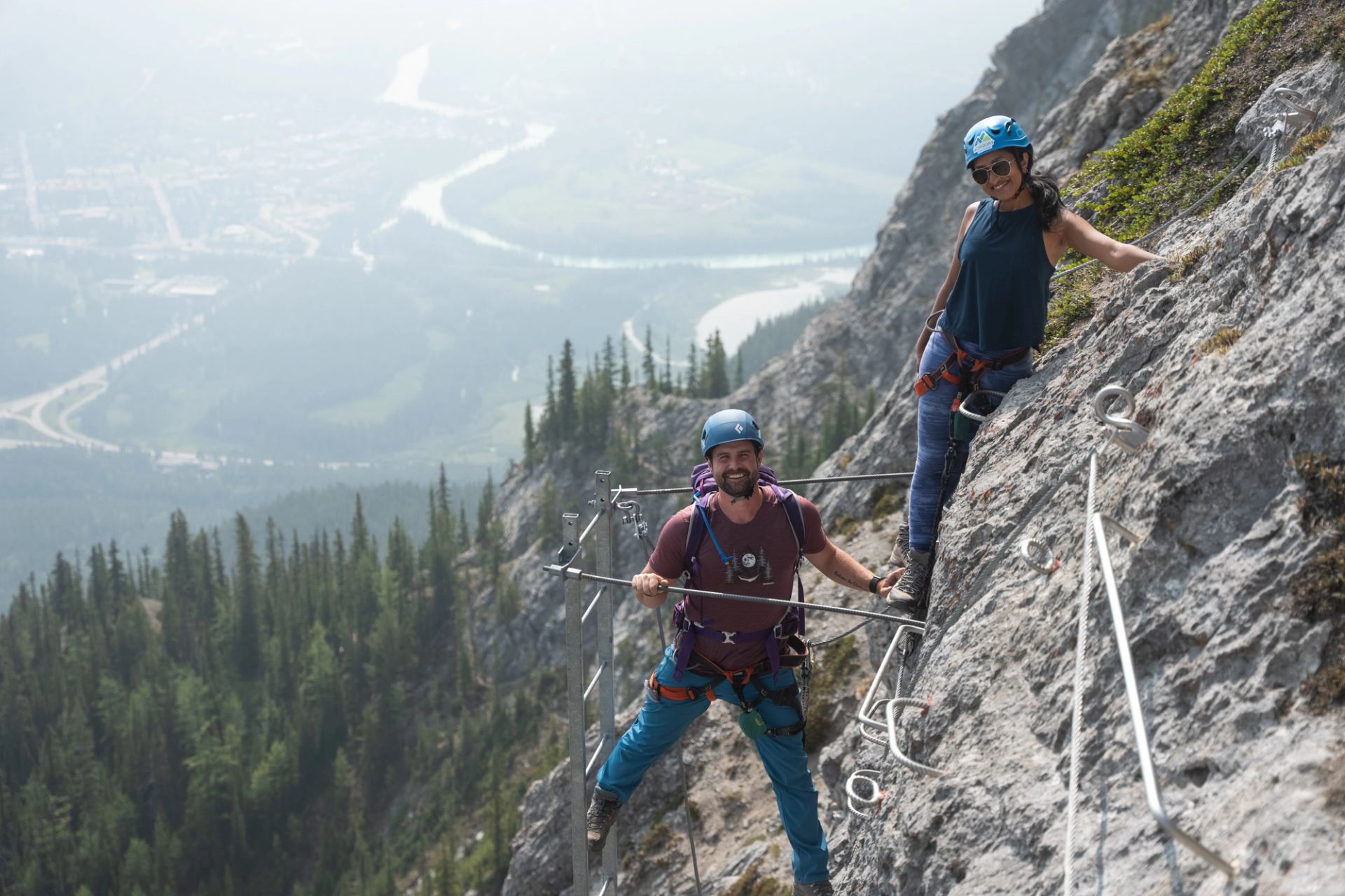 Two climbers on rocky mountain with gear, overlooking a scenic valley.