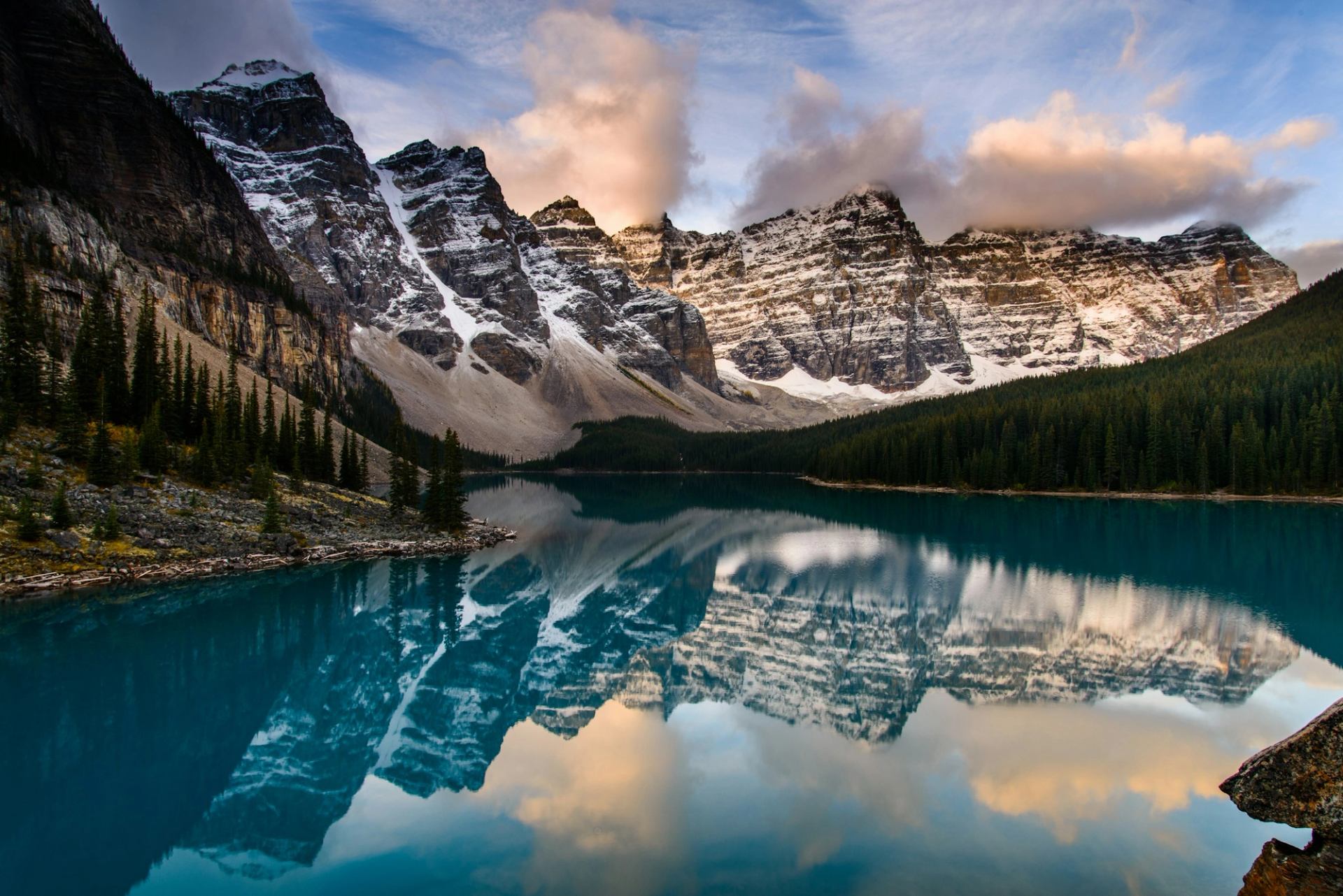 Snow-capped mountain range reflected in calm turquoise lake, bordered by dense evergreen forest.